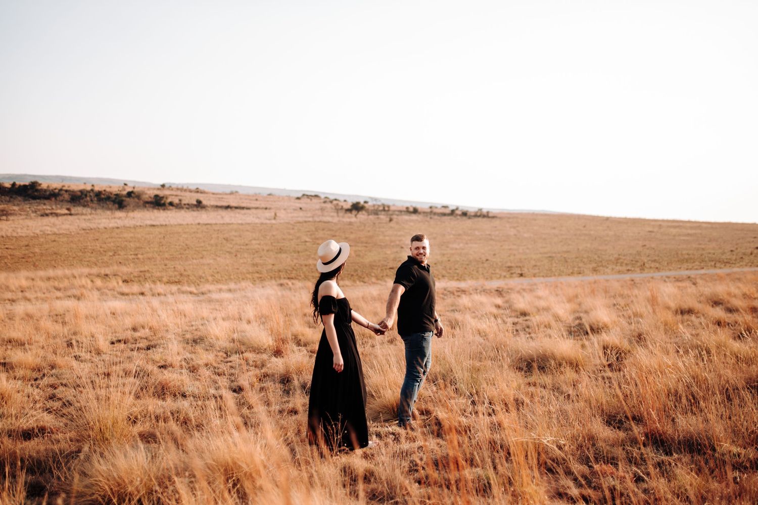 Two people walk hand in hand through golden autumn grass on a rolling hillside at sunset.
