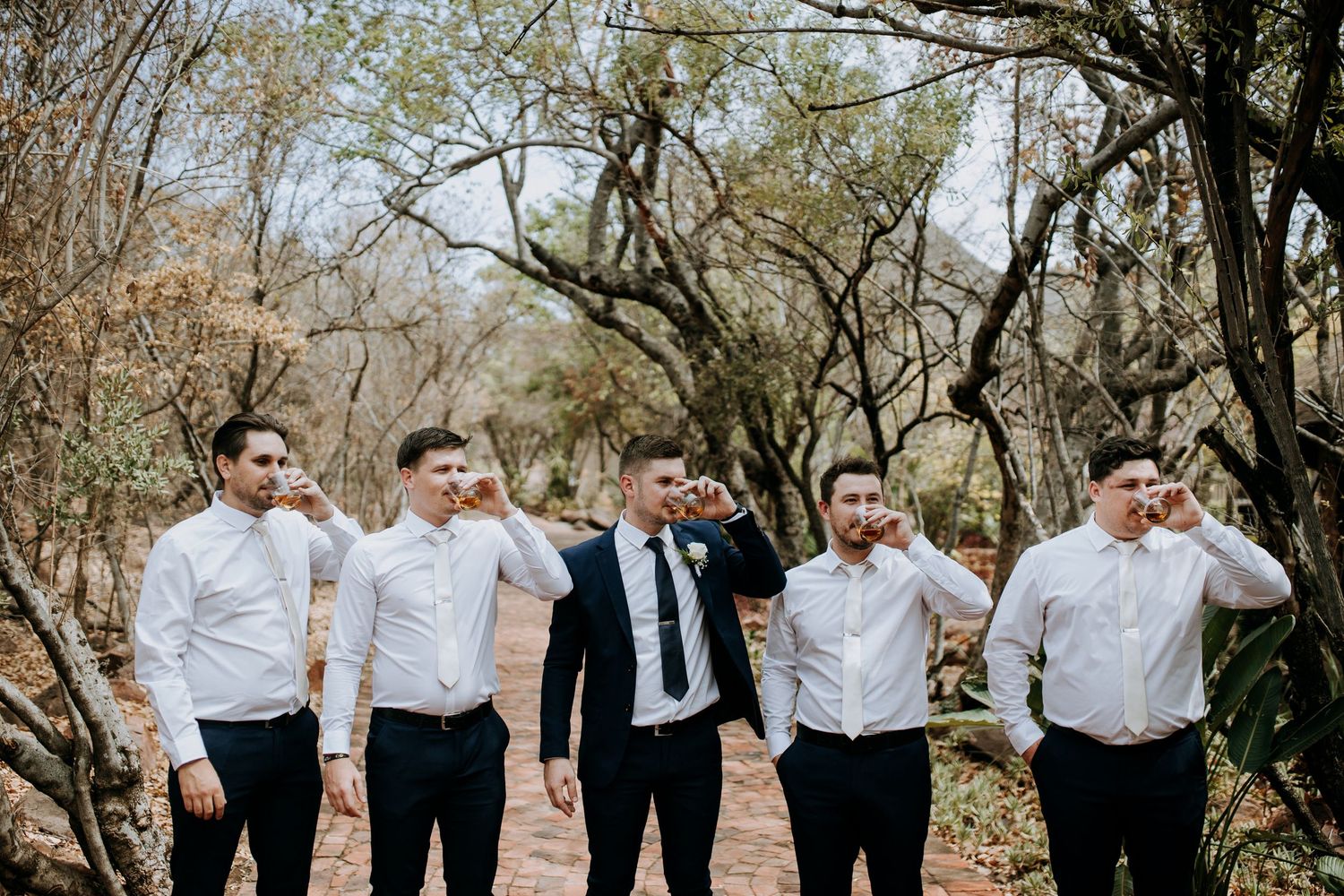 A joyful group in coordinated black and white attire stands together outdoors in a natural setting with autumn trees.