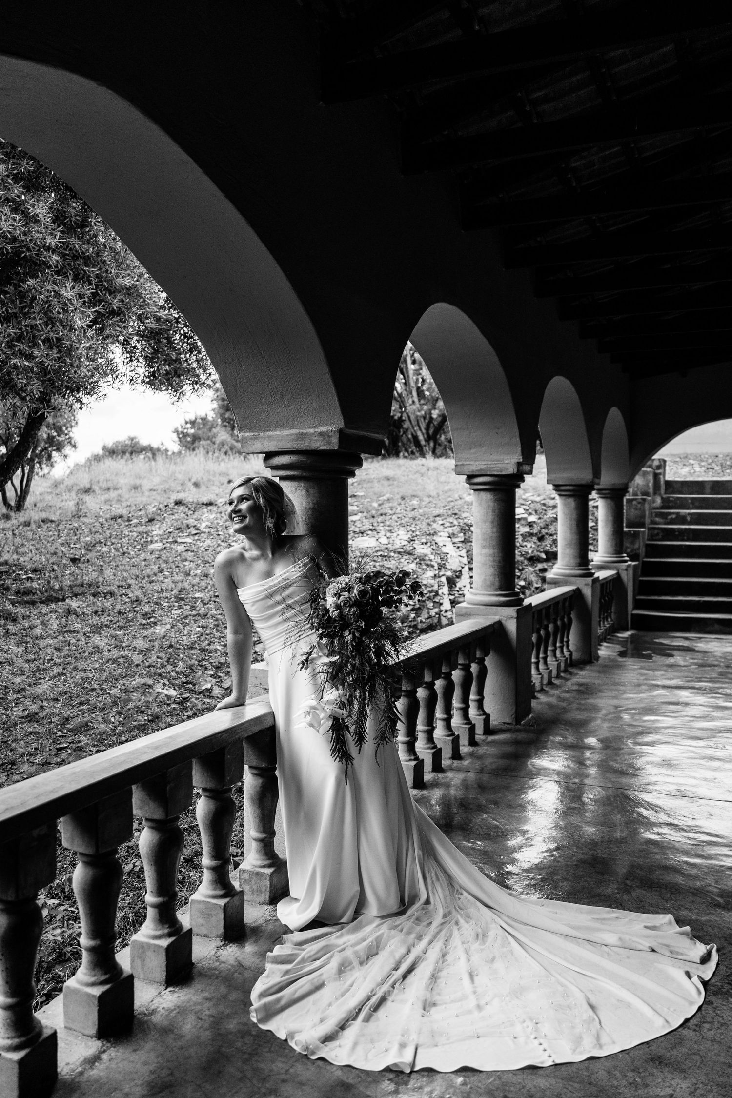 A flowing white wedding dress train cascades elegantly beneath arched columns in a dramatic black and white composition.