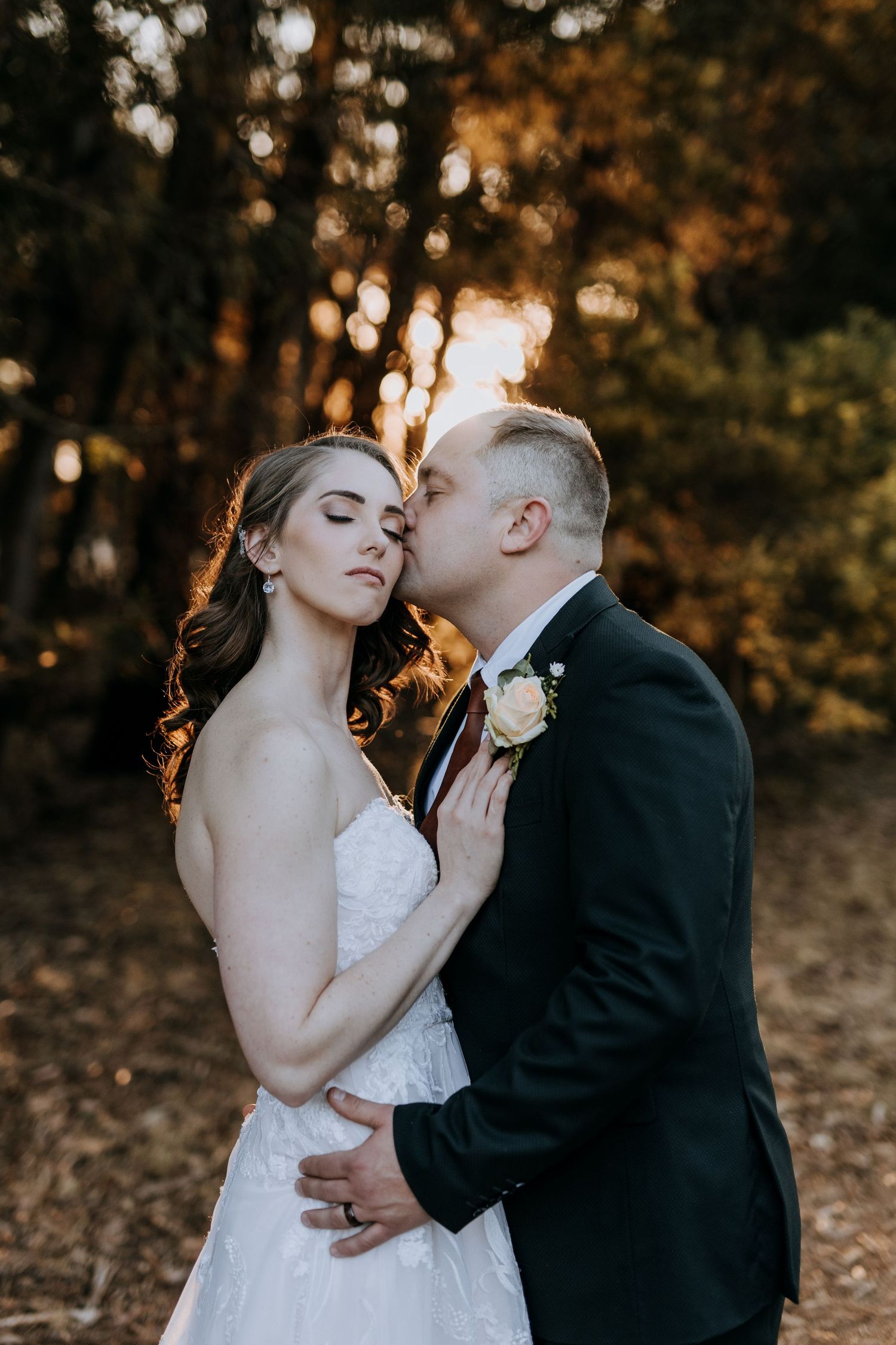 A romantic couple shares an intimate moment during their wedding photoshoot in a golden-lit forest setting.