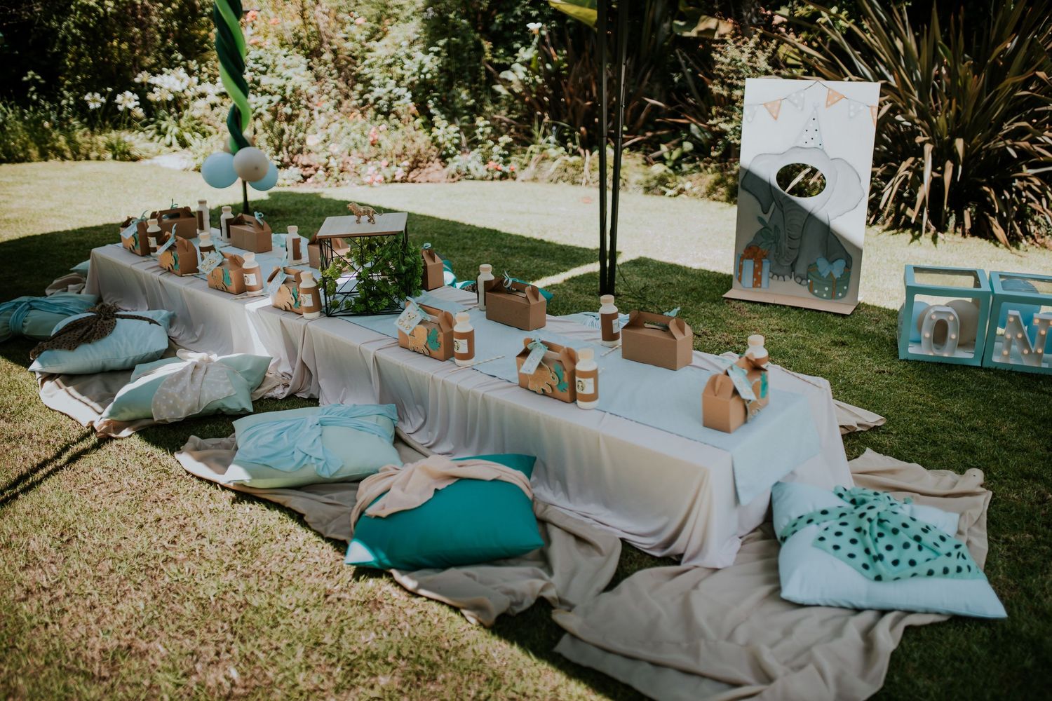 Outdoor picnic setup with floor cushions, white tablecloth, and turquoise accents in a sunny garden setting.