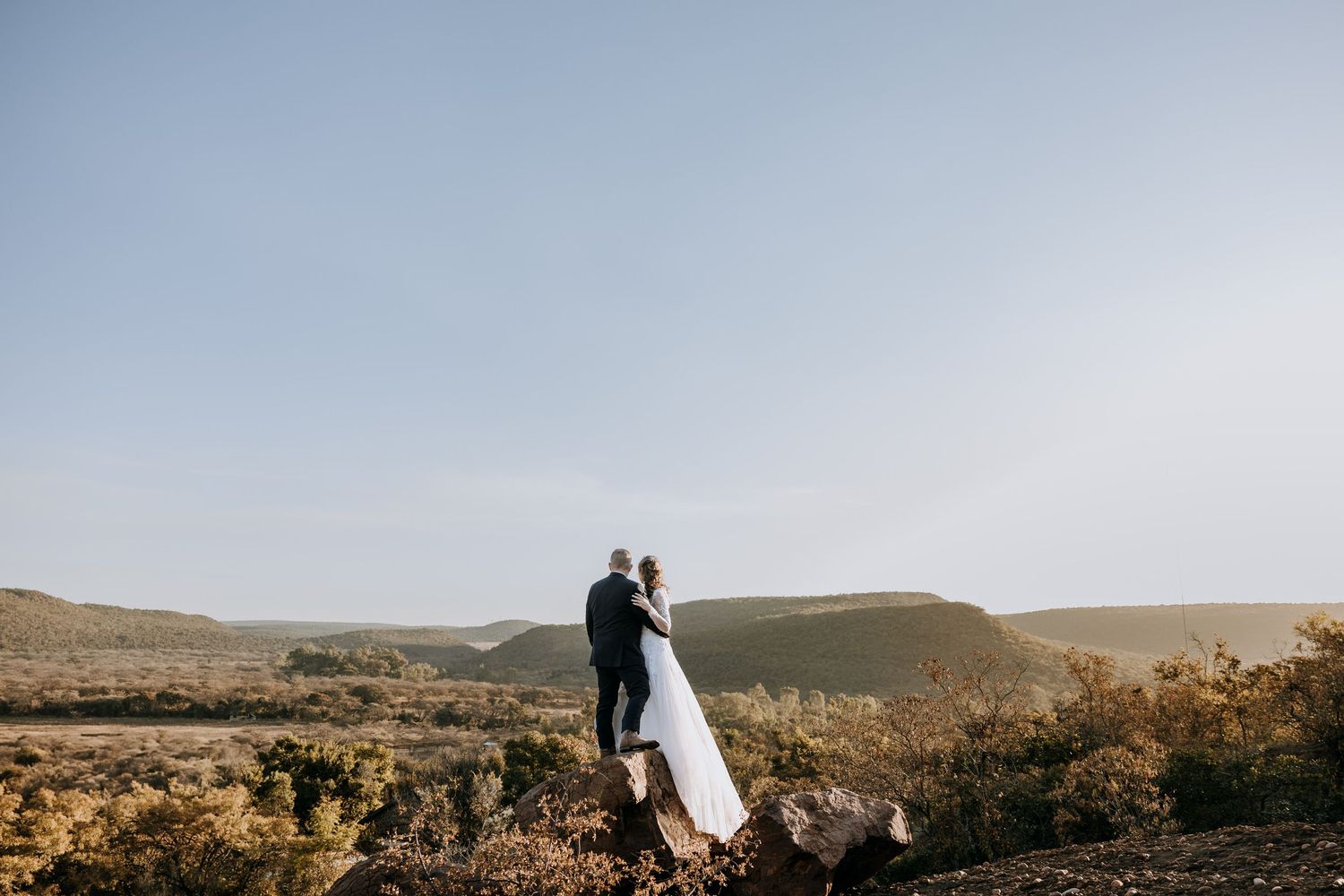 Romantic wedding couple stands together on mountain ridge during sunset in desert landscape.