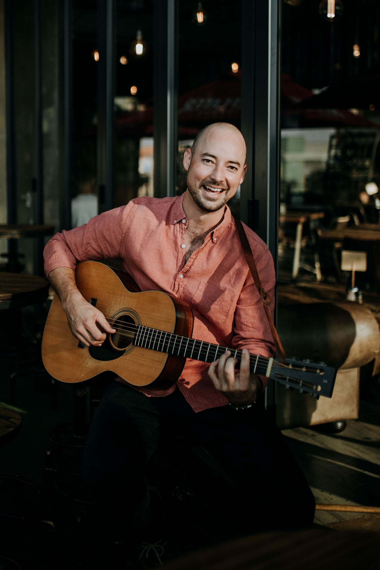 A person in a pink button-up shirt plays acoustic guitar while sitting in a dimly lit restaurant setting with hanging lights.