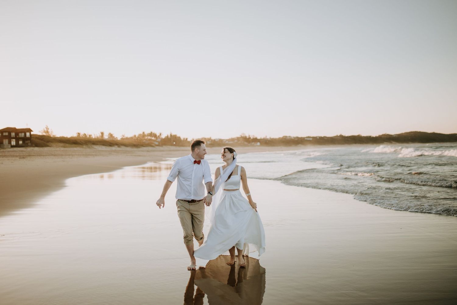 Couple strolls hand in hand along a peaceful beach shoreline at sunset with light reflecting off the wet sand.