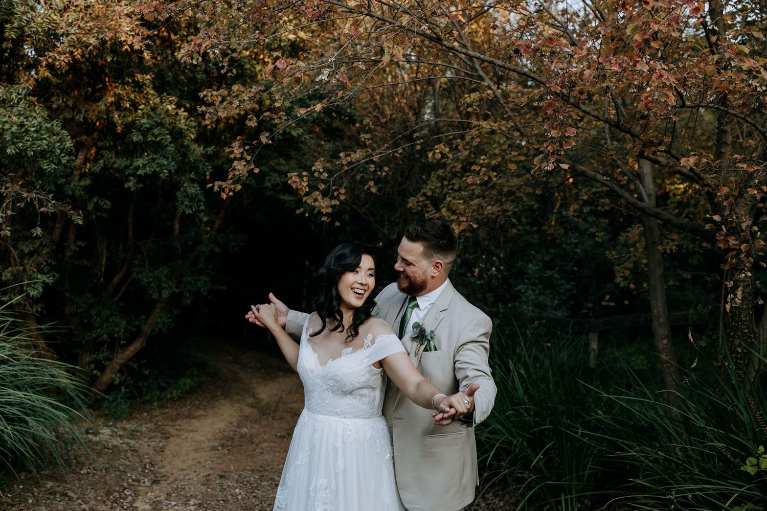A couple in wedding attire walks hand in hand through a wooded path during autumn.