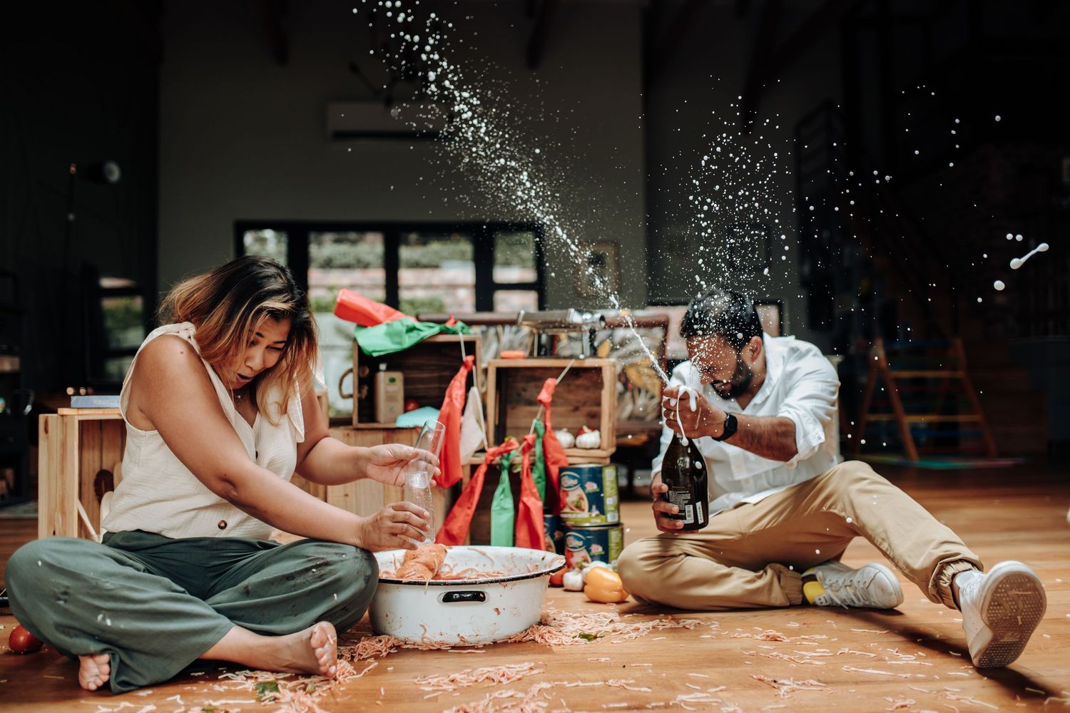 Two people sit on a wooden floor surrounded by art supplies and paper while collaborating on a craft project.