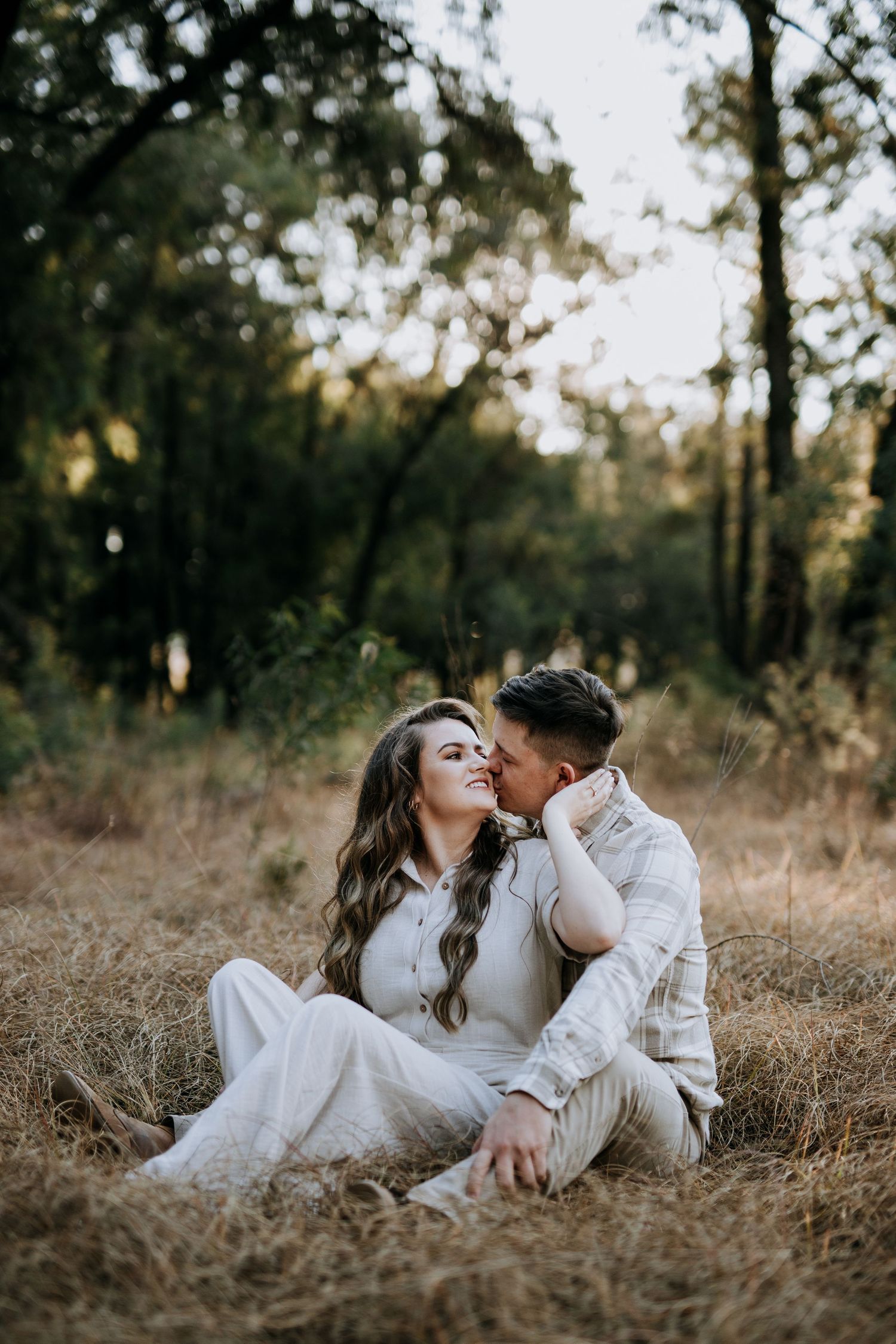 A couple in white clothing shares intimate moments while laying in a golden field during sunset.