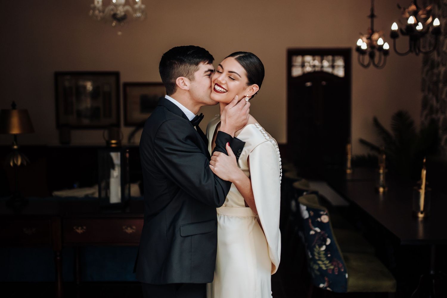 A romantic couple shares an intimate kiss in an elegant ballroom setting with sparkling chandeliers overhead.