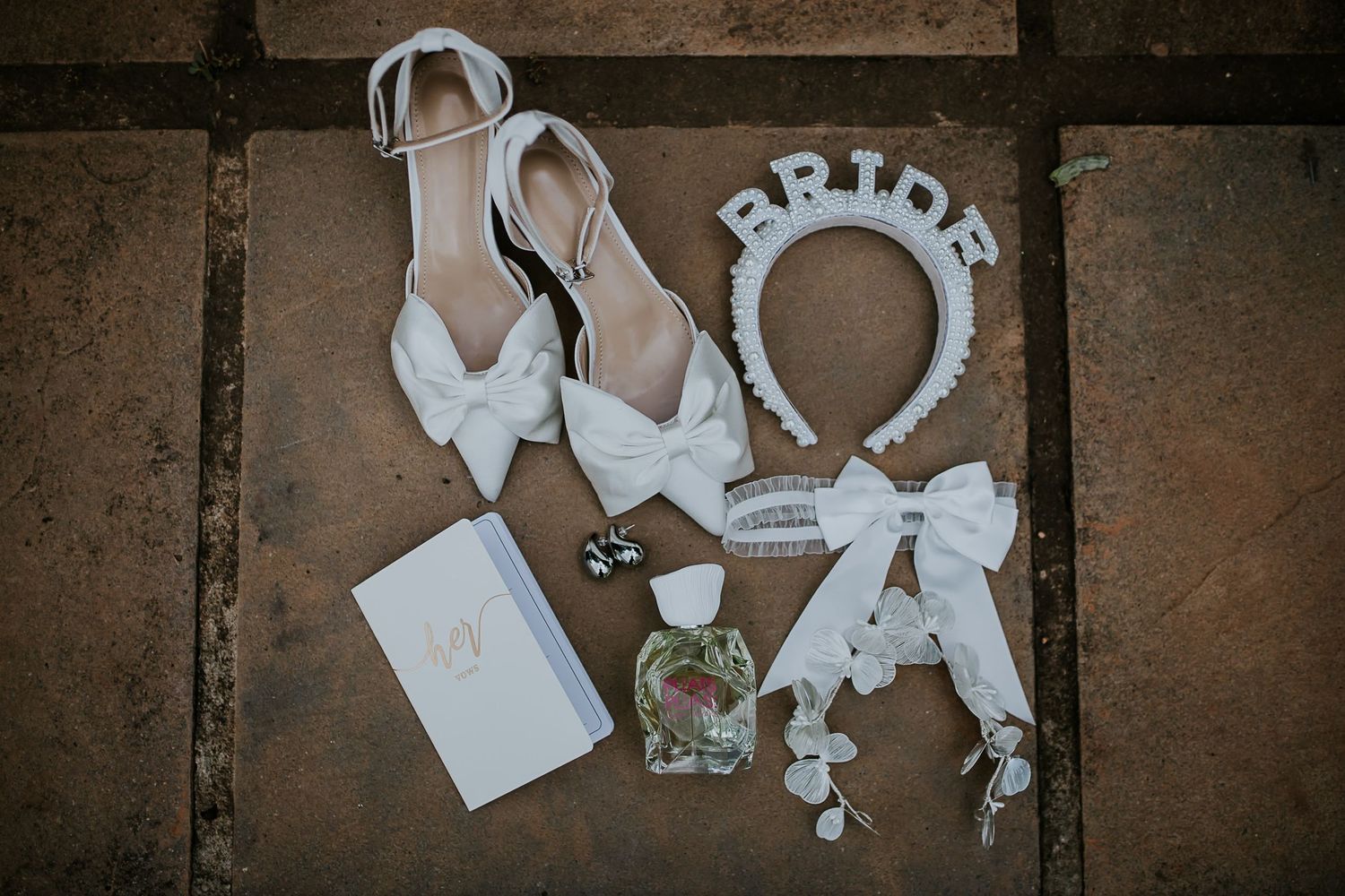 Wedding accessories including white bow shoes, bride headband, and delicate decorative items arranged on stone surface.
