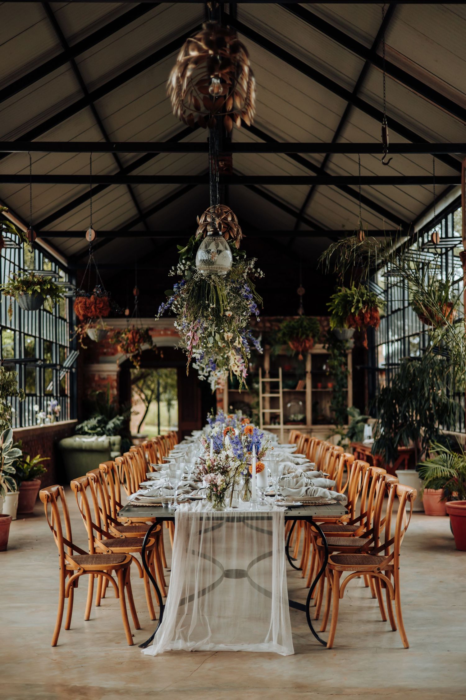 Wedding reception table setup with wooden chairs in a greenhouse-style venue featuring elegant floral arrangements.