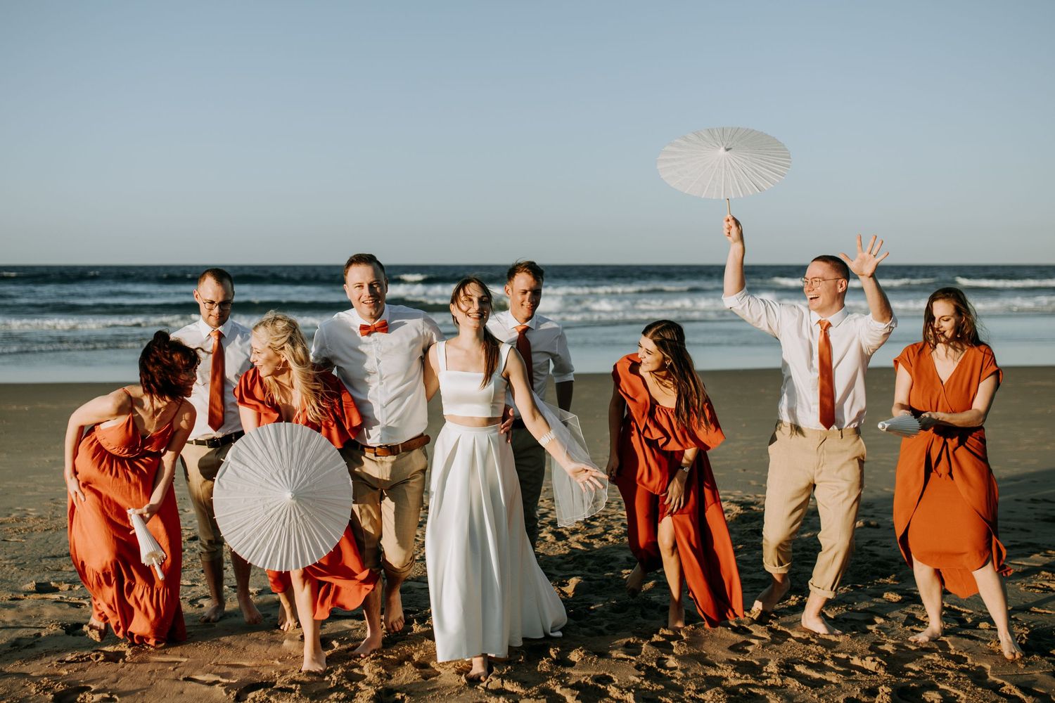 Wedding party in coral dresses and white gowns poses playfully along scenic beach at sunset.