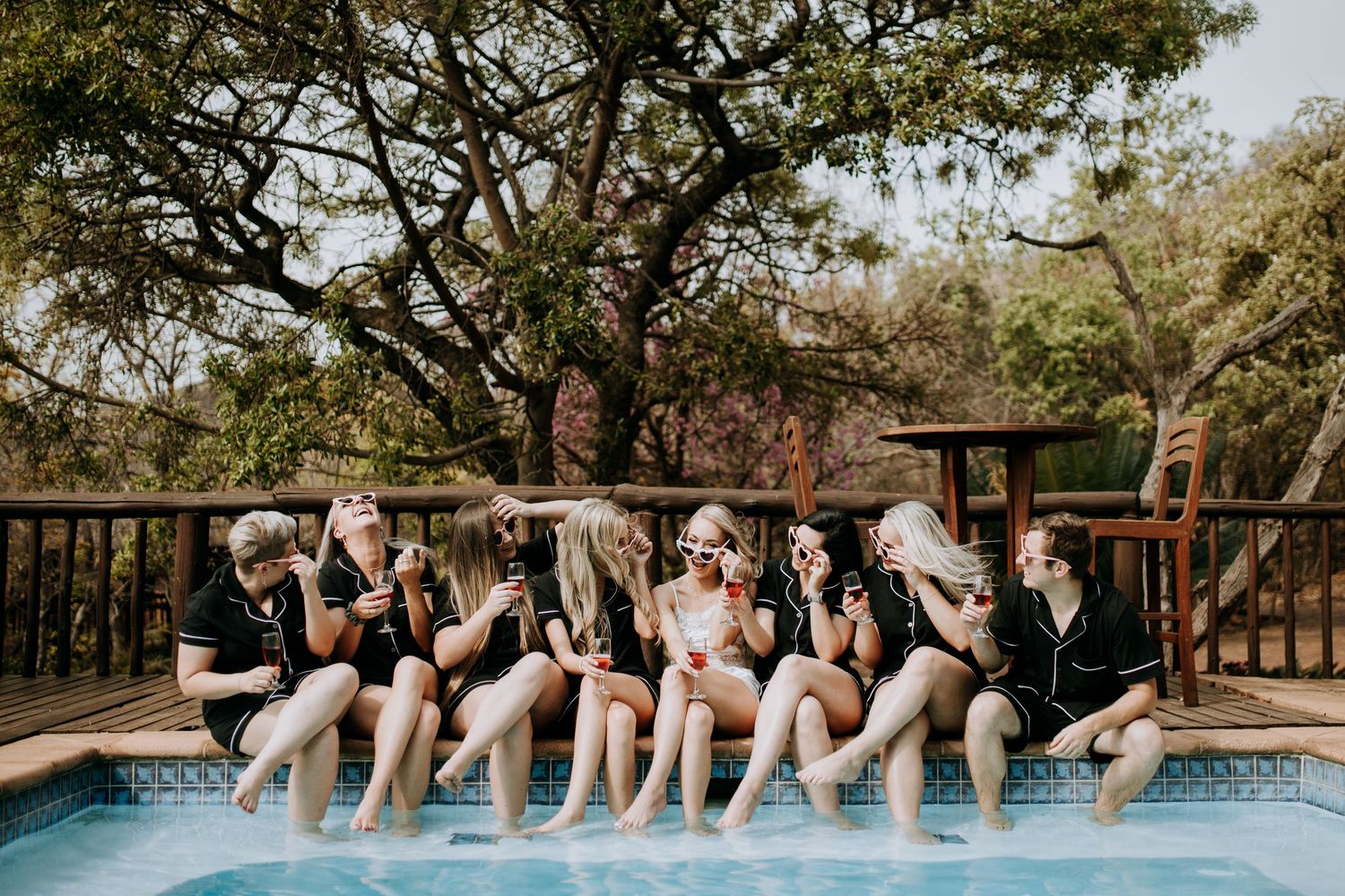 Wedding party lined up on wooden deck near pool doing synchronized jumping pose during golden hour sunset.