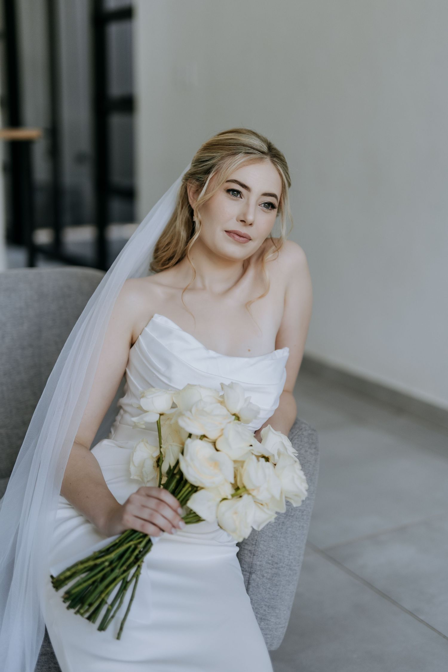 A bride in a classic white wedding dress holds an elegant bouquet of white flowers against a light gray backdrop.