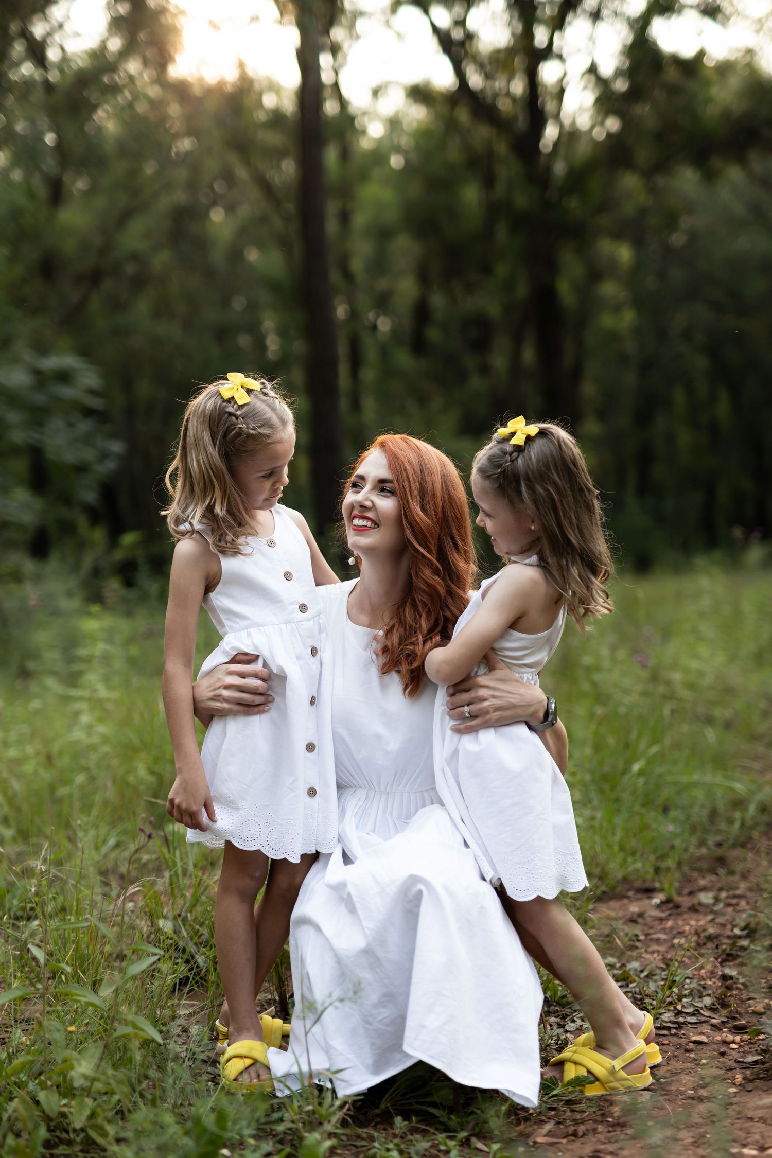 Three friends in white dresses share joyful moments together on a wooded path during golden hour.
