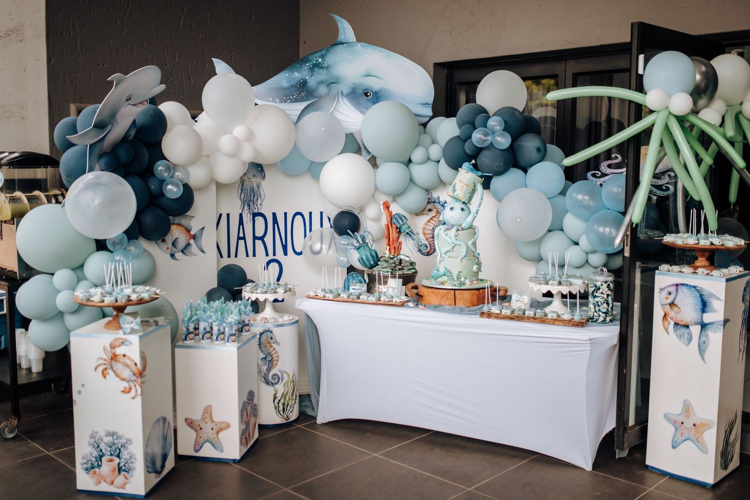 Elegant baby shower party setup with blue and white balloon garlands and themed decorative boxes along the wall.