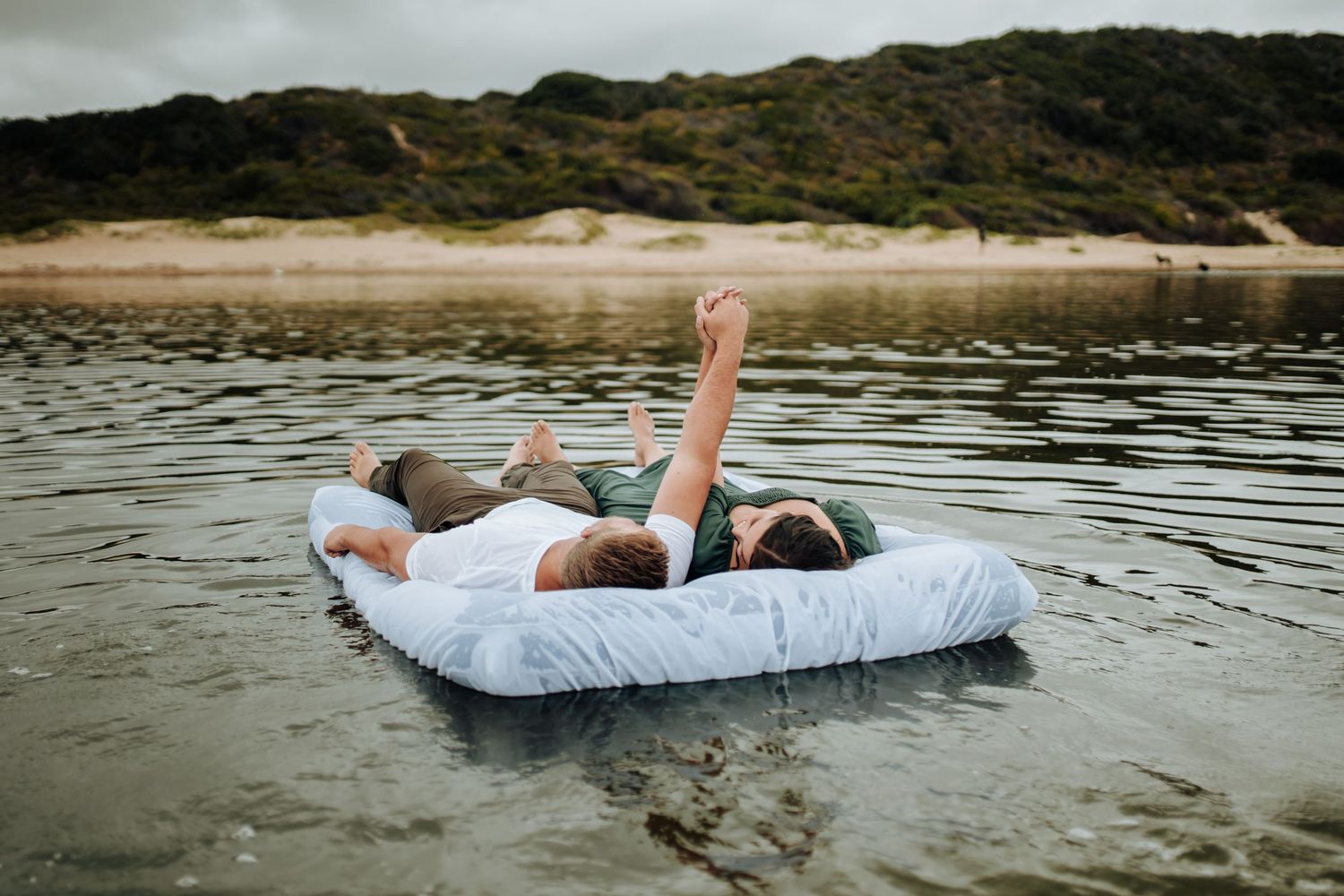 Two people relax on a white inflatable raft floating on calm waters near a sandy beach.