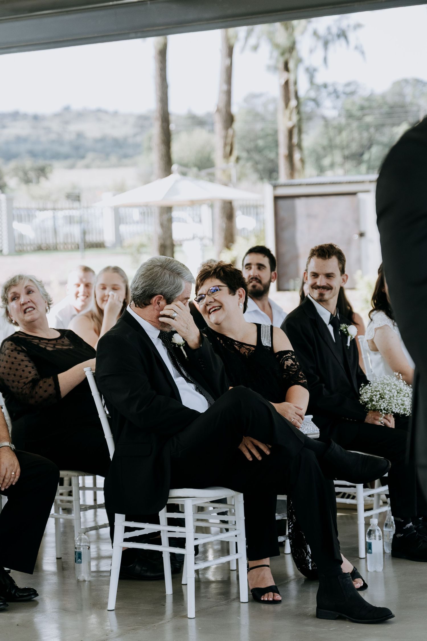 Wedding guests laugh and enjoy themselves while seated in white chairs at an outdoor reception.