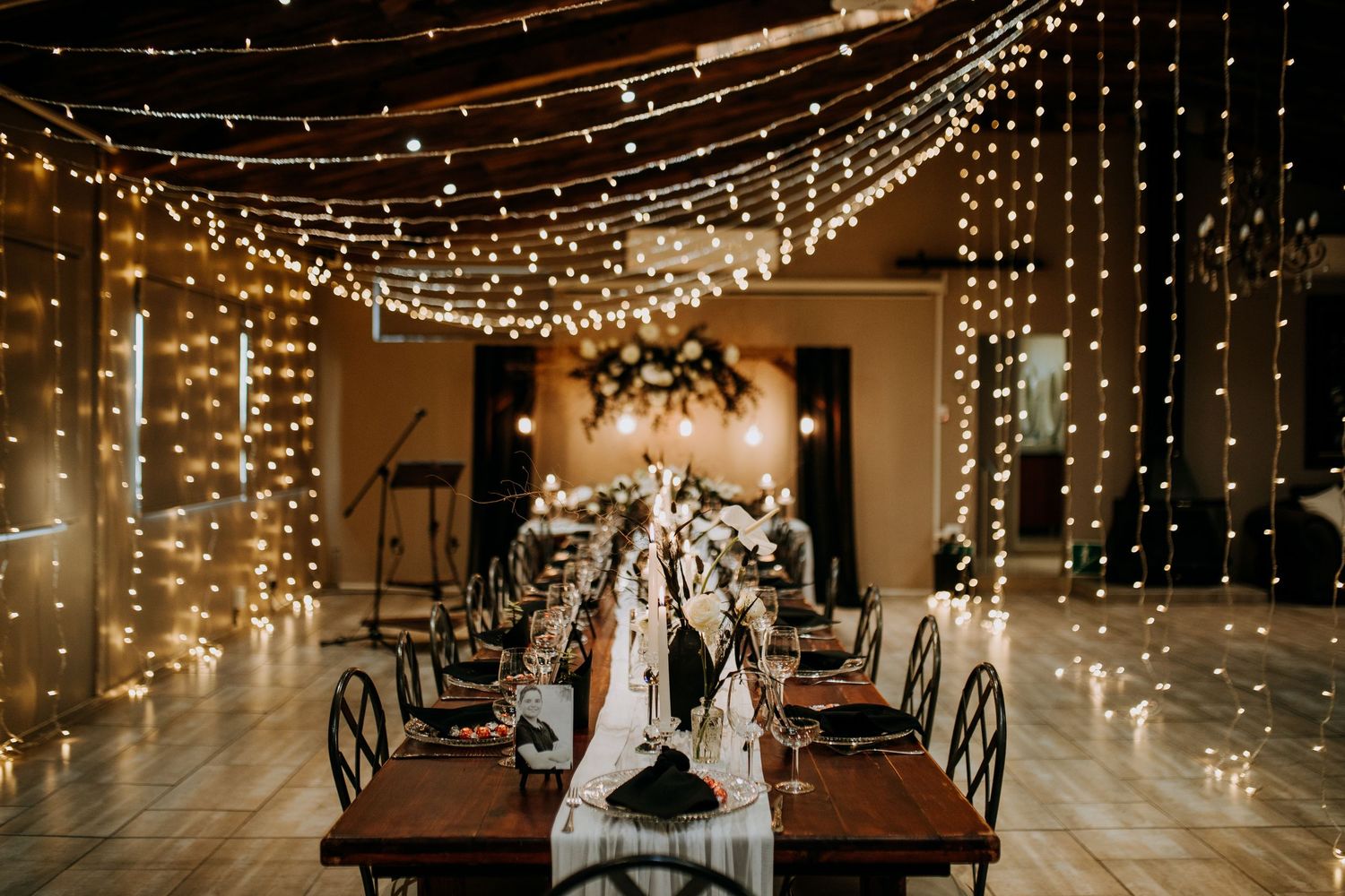 String lights create a romantic canopy over dark wooden tables in an elegant reception venue setting.