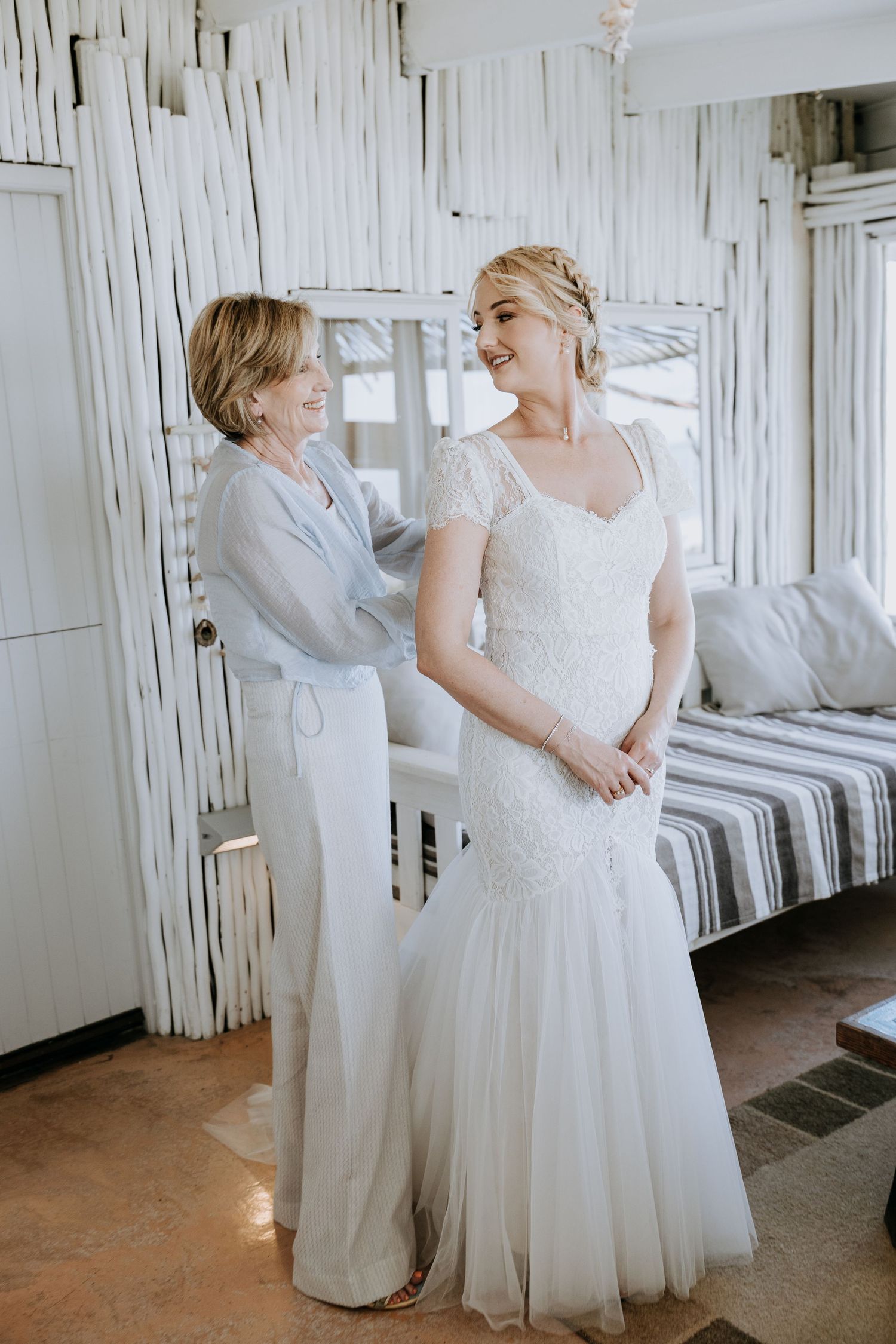 Two people wearing white formal dresses stand close together in a bright, airy room with white curtains.