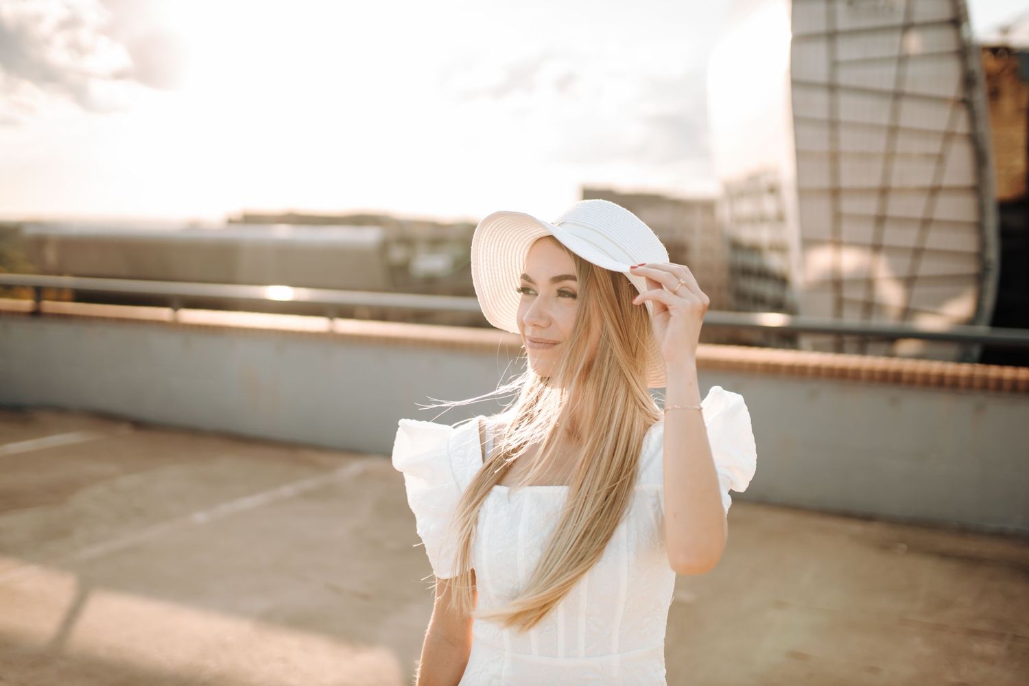 Woman in a white sun hat and dress sits gracefully on wooden steps in warm sunlight.