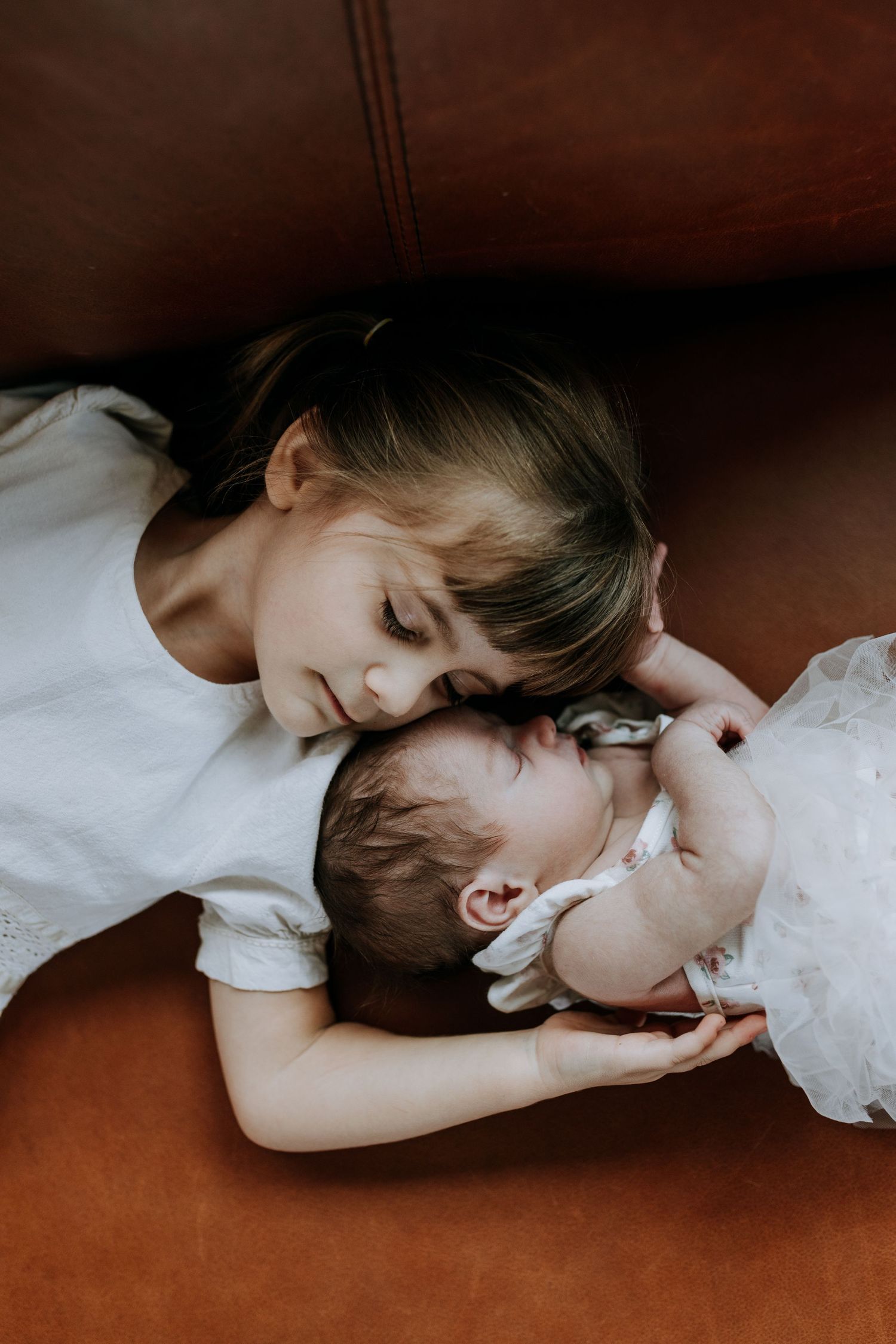 Older girl in white dress tenderly cuddling sleeping newborn baby sibling on brown leather sofa.