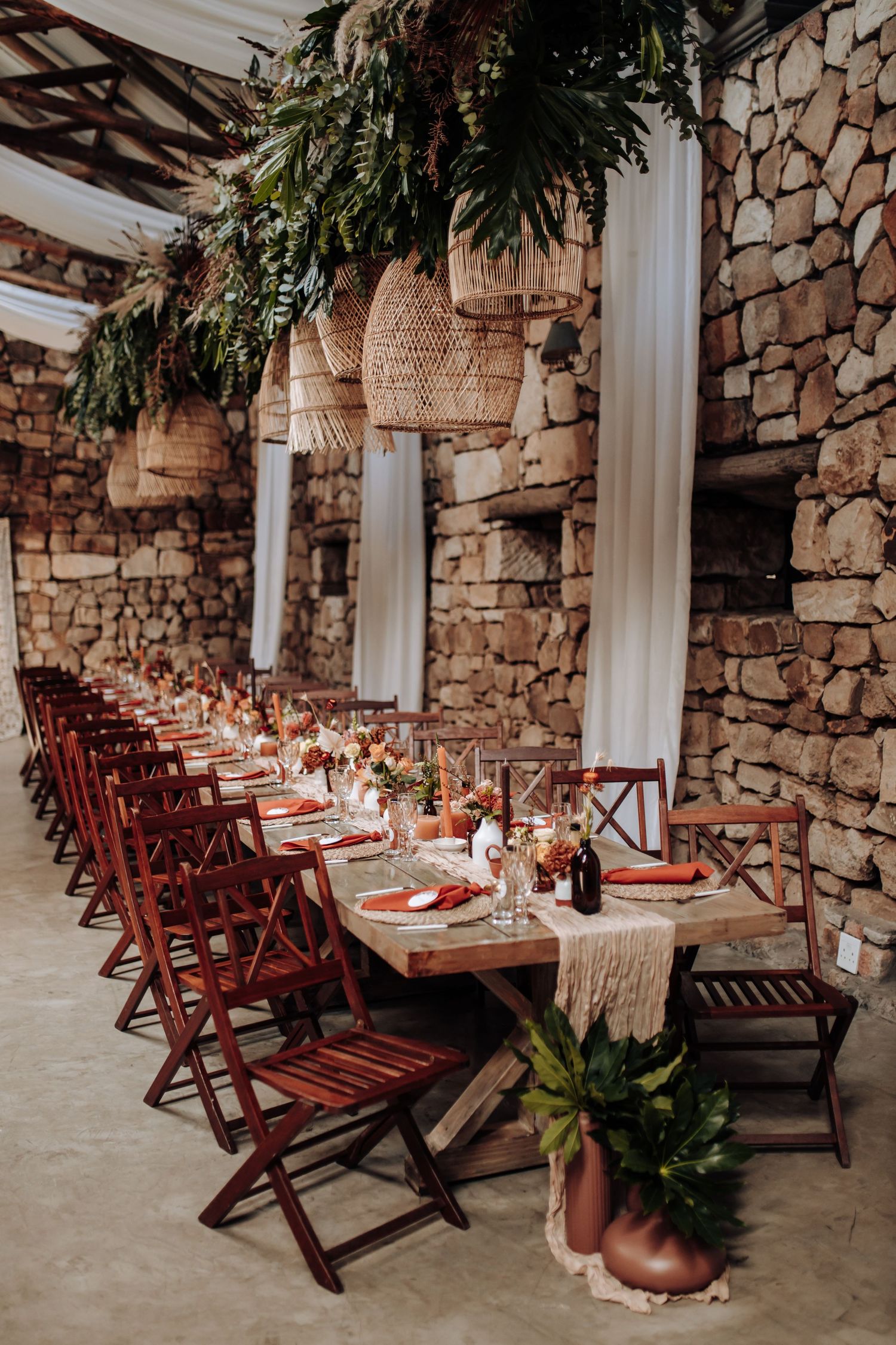 Rustic wedding reception setup with long wooden tables and dark red chairs against natural stone walls.