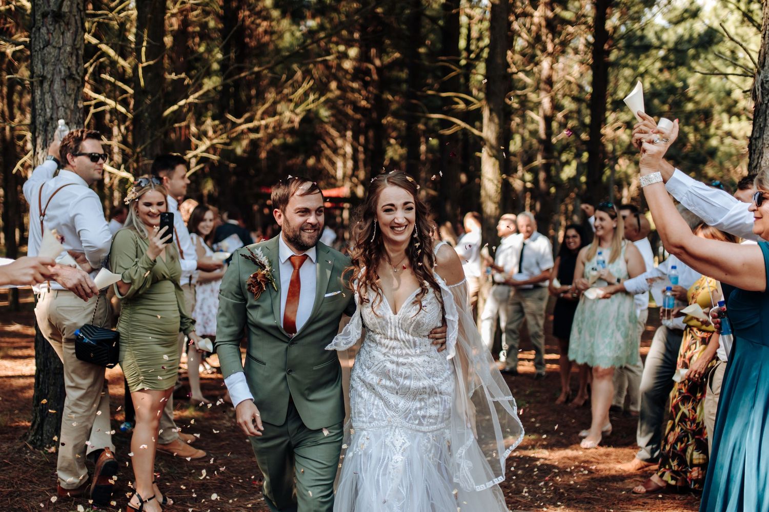 A wedding celebration takes place in a sun-dappled forest with guests dressed in formal attire cheering along a wooded path.
