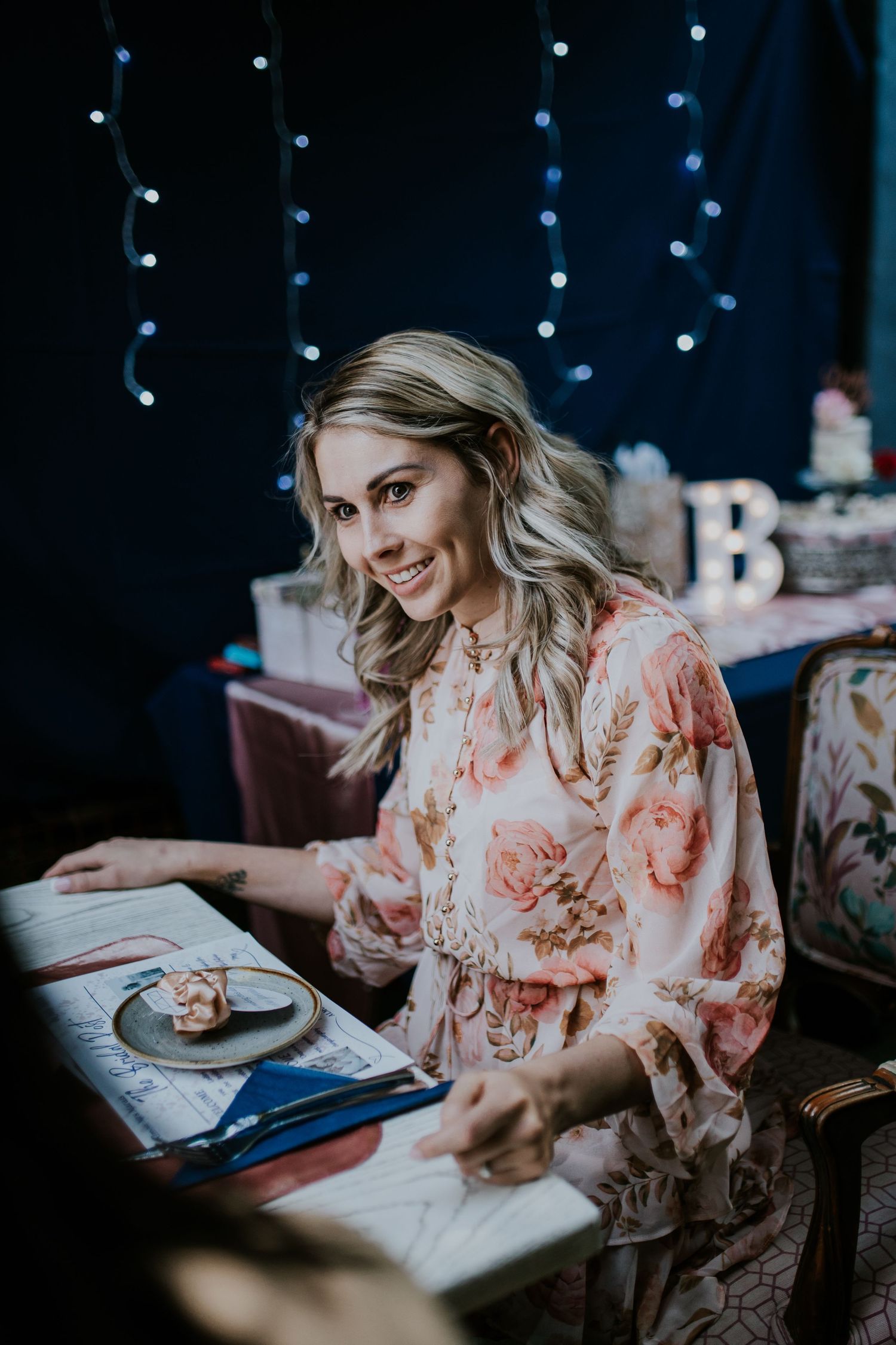 Person in floral dress sits at elegant table with string lights above in dark atmospheric setting.