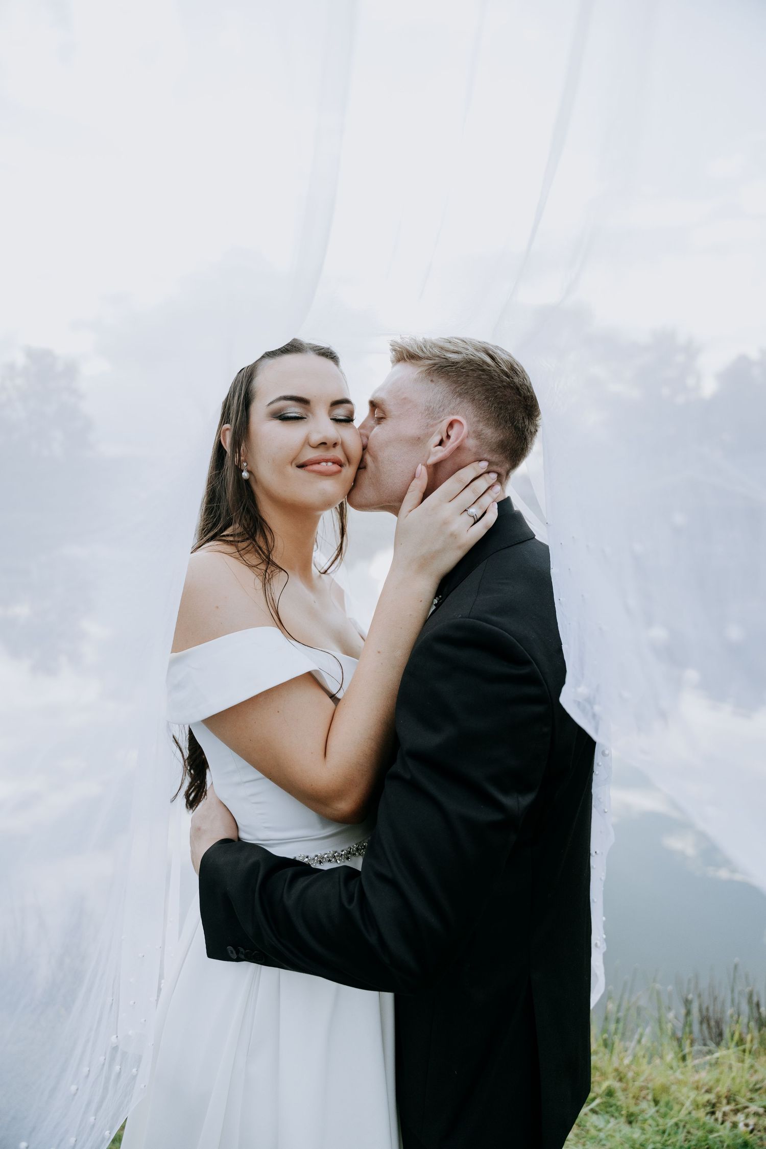 A tender wedding moment captured against a mountain backdrop with soft white veil flowing in the wind.