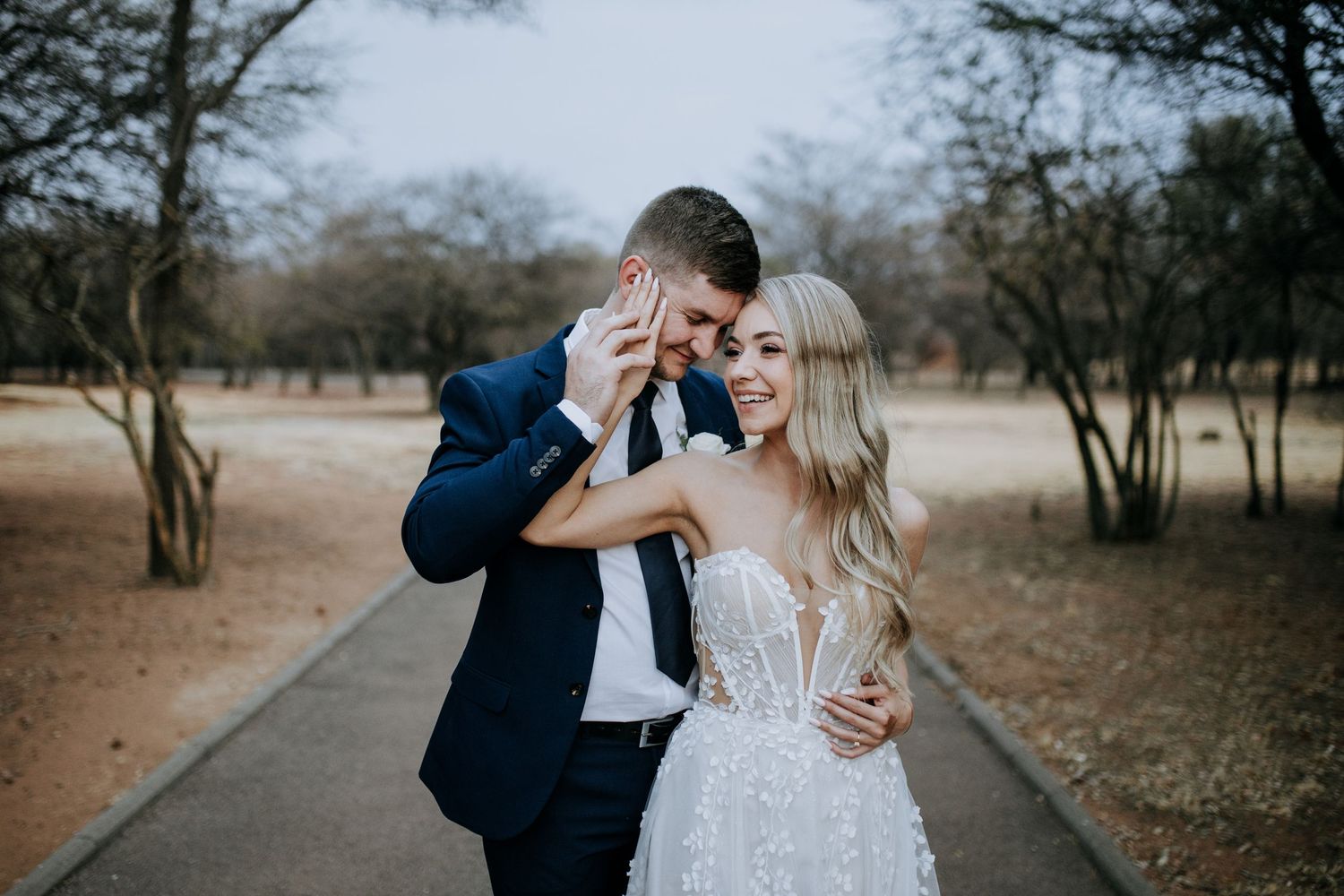 A bride in a detailed white gown and groom in navy suit share an intimate moment on a tree-lined path.