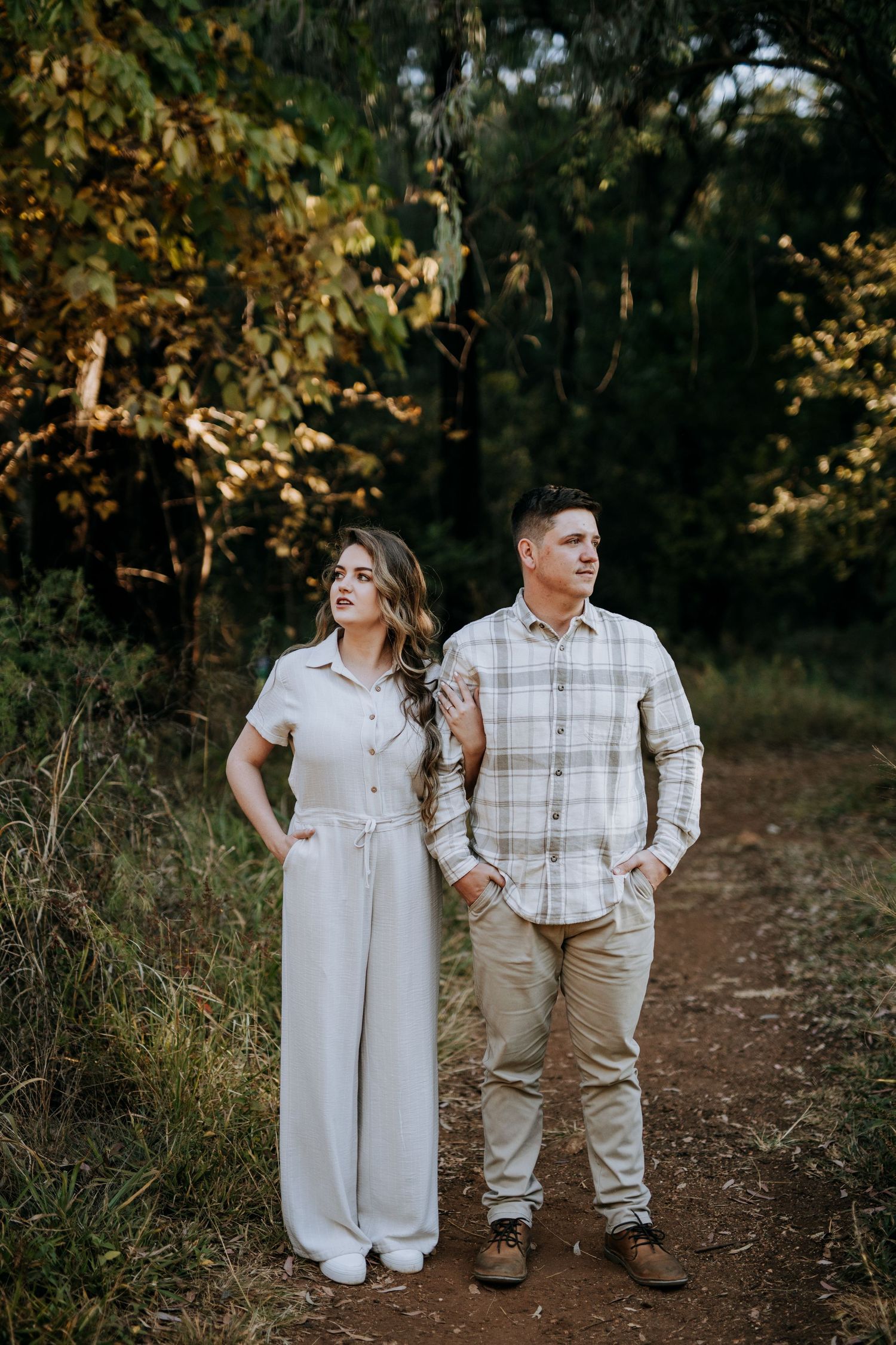 A couple poses together on a woodland trail wearing coordinated casual outfits in neutral tones.