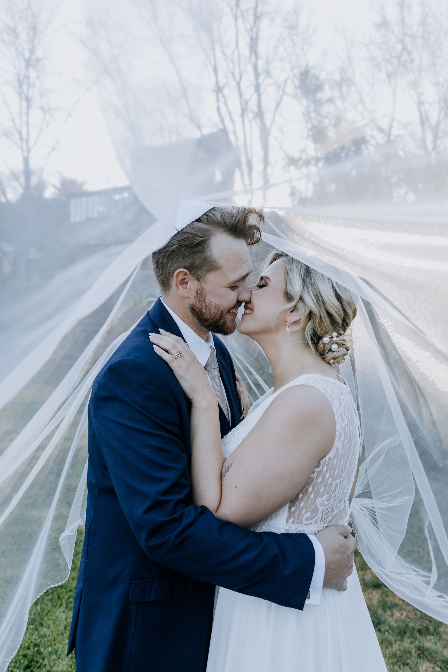 Wedding couple shares an intimate kiss under a flowing veil while surrounded by misty forest scenery.