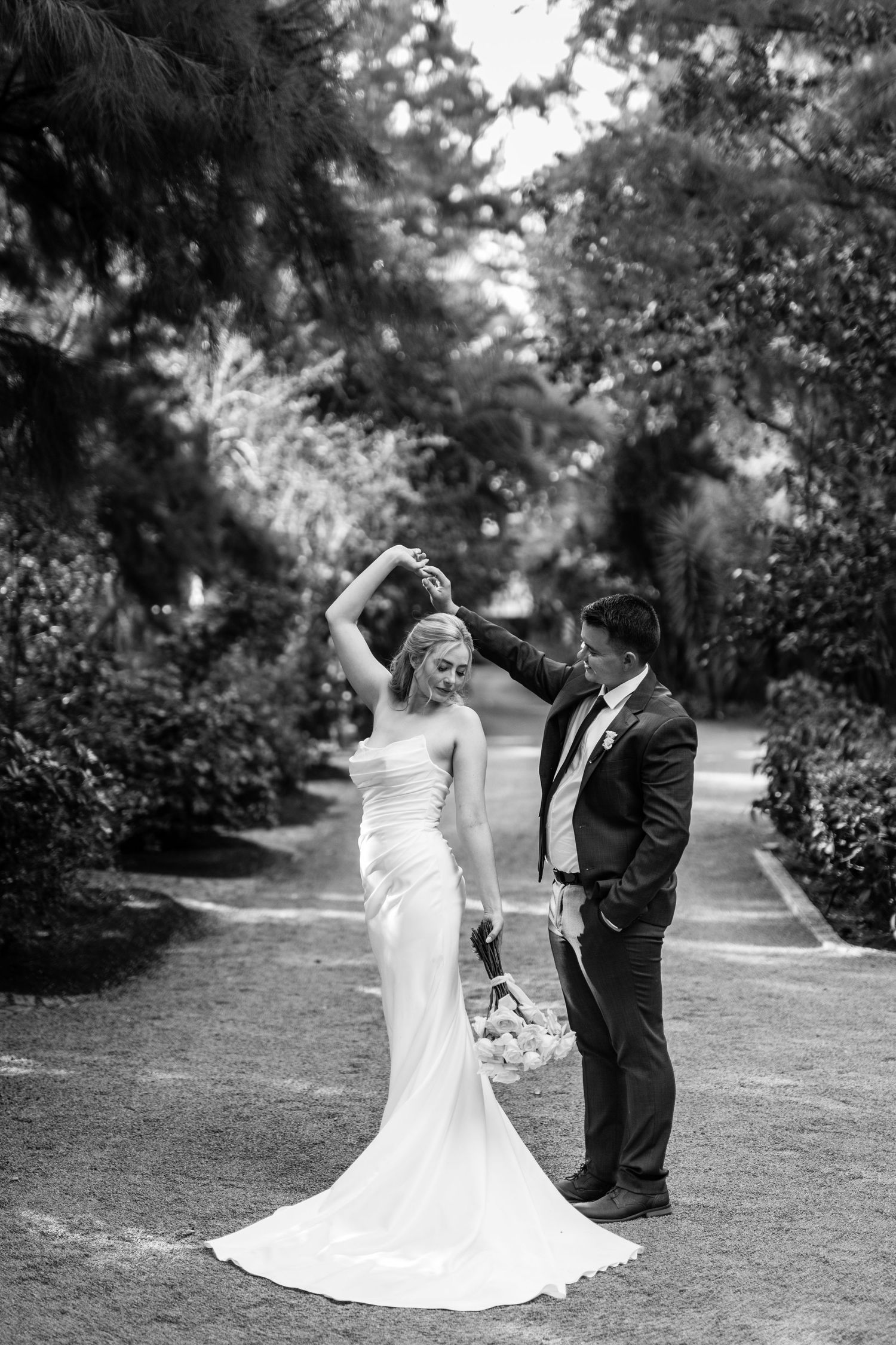 A romantic black and white wedding photo of a couple dancing together on a garden path surrounded by lush greenery.