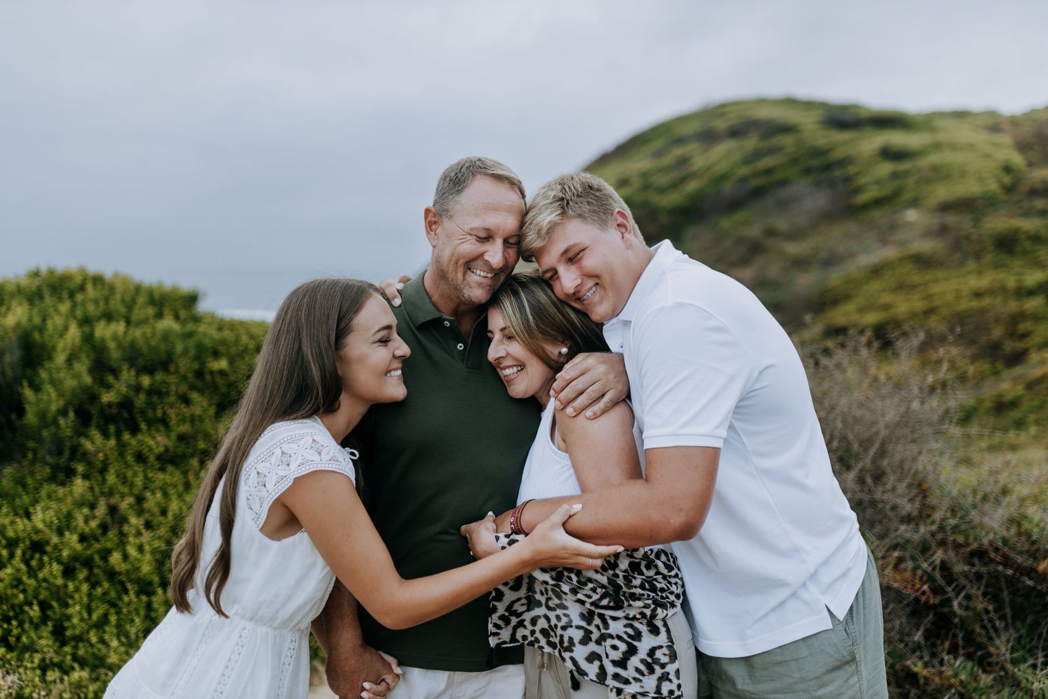 A family shares a joyful moment together outdoors on a green hillside against a cloudy sky.
