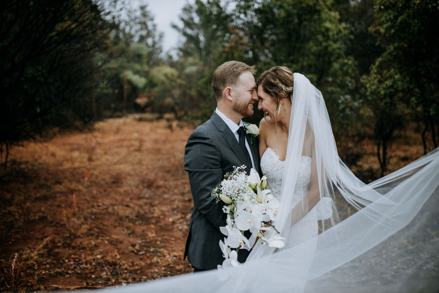 A romantic wedding couple embraces on a forest path while a long flowing veil catches the breeze.