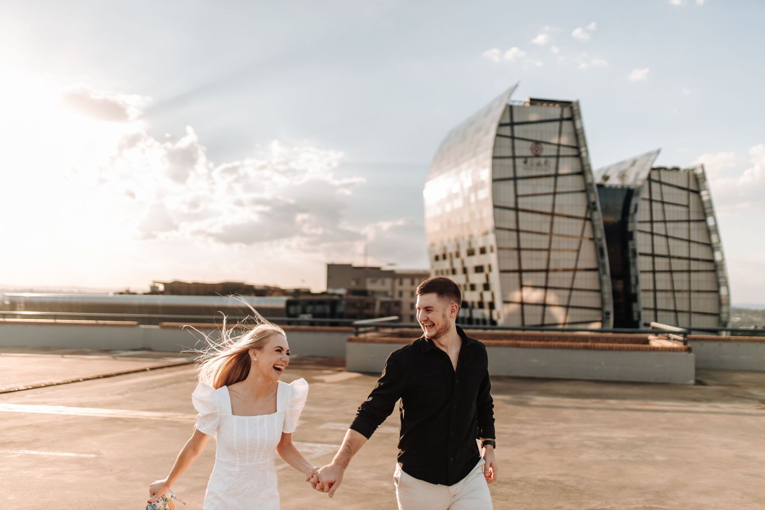 A couple holds hands while walking on a rooftop plaza with modern glass buildings in the background at sunset.