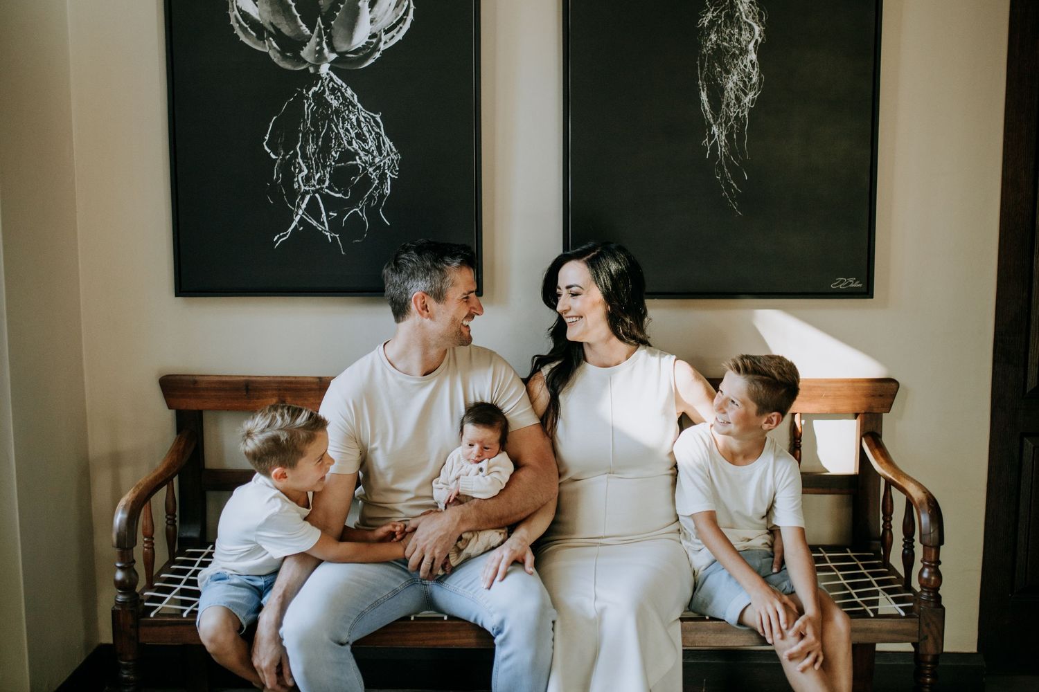 A family dressed in white sits together on a bench, sharing smiles and laughter inside a room with artistic wall decor.