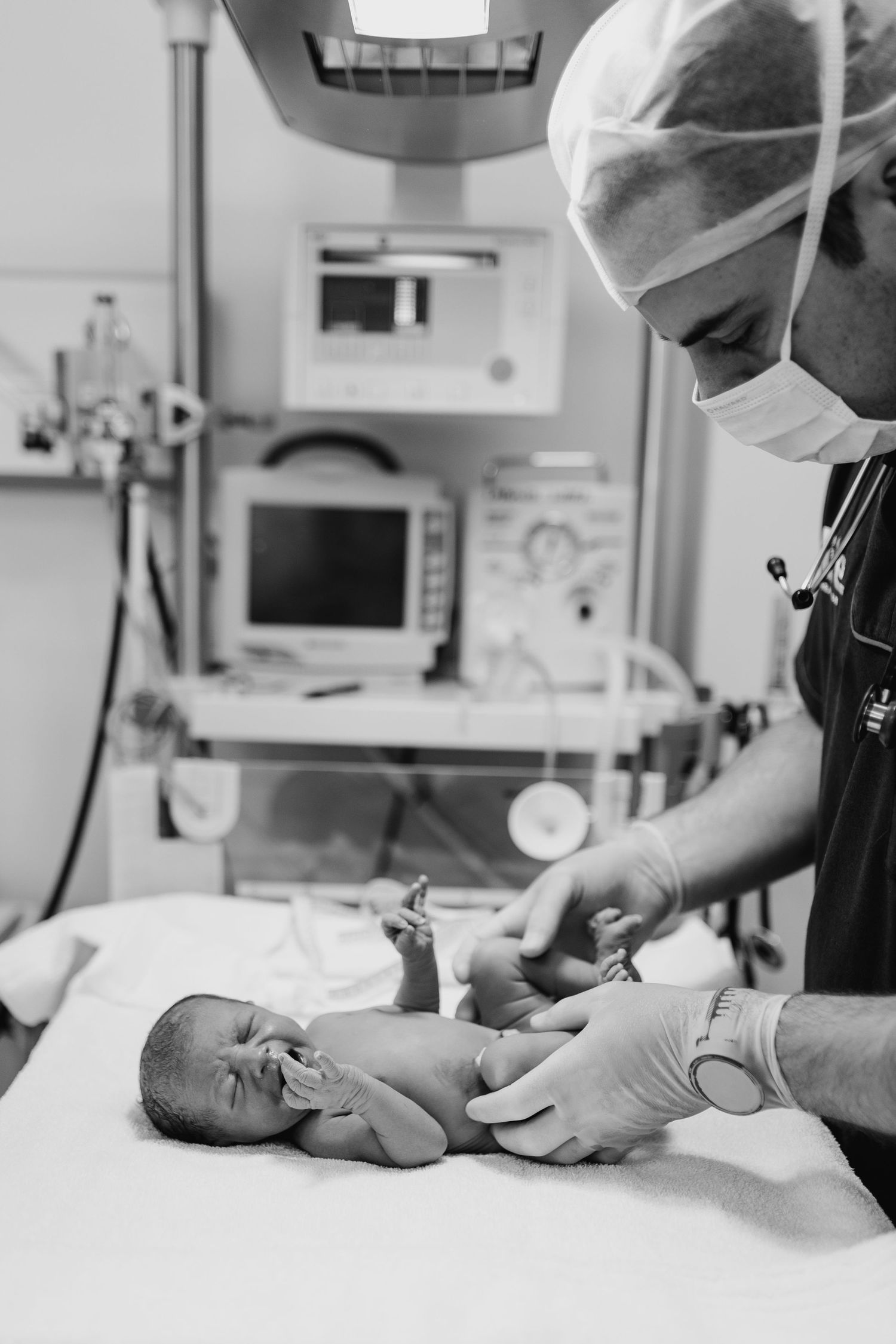 Black and white photo of a doctor in surgical mask and gloves examining a newborn baby in a hospital delivery room.