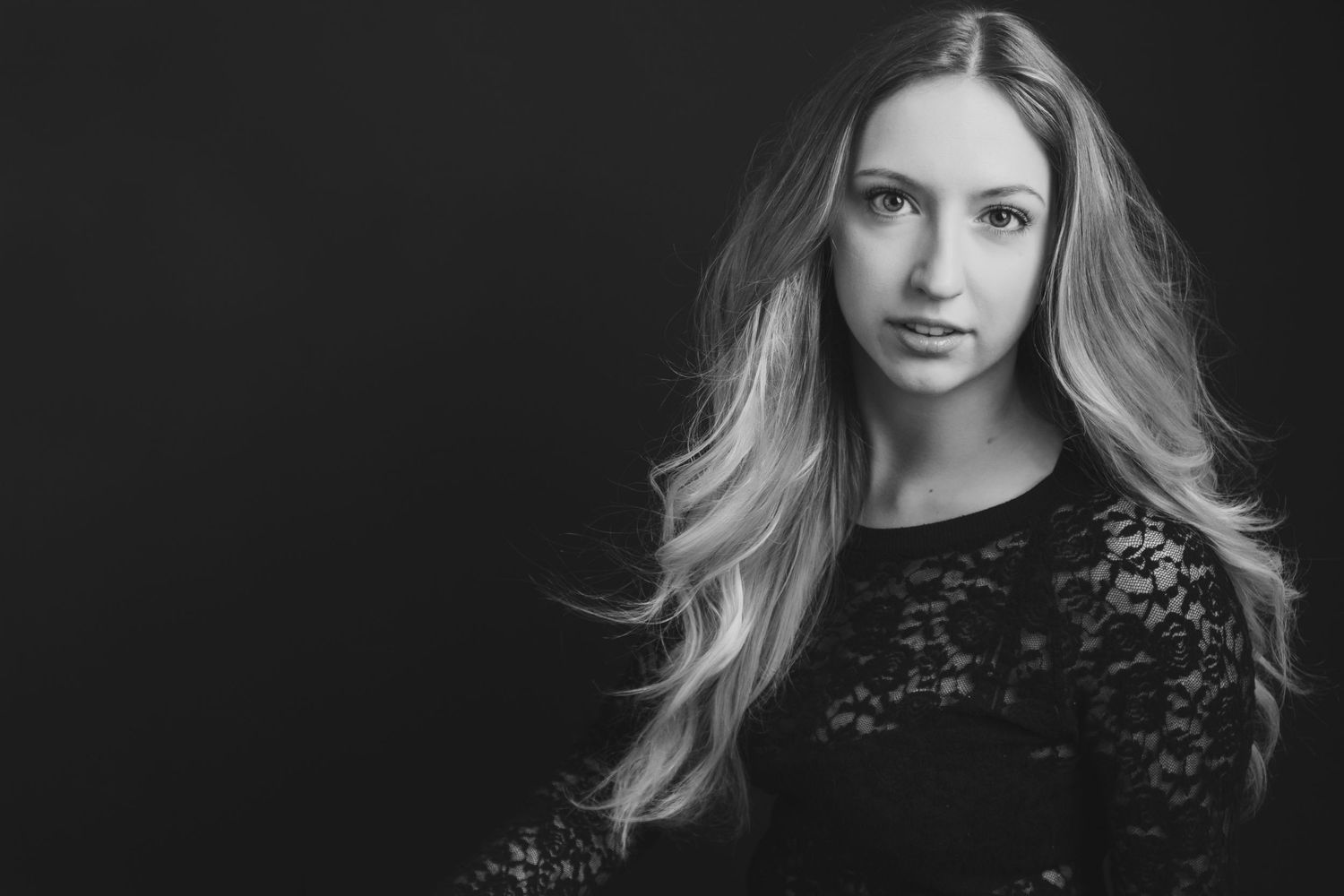 Black and white portrait of a young blonde woman in lace top, gazing at camera against dark background.