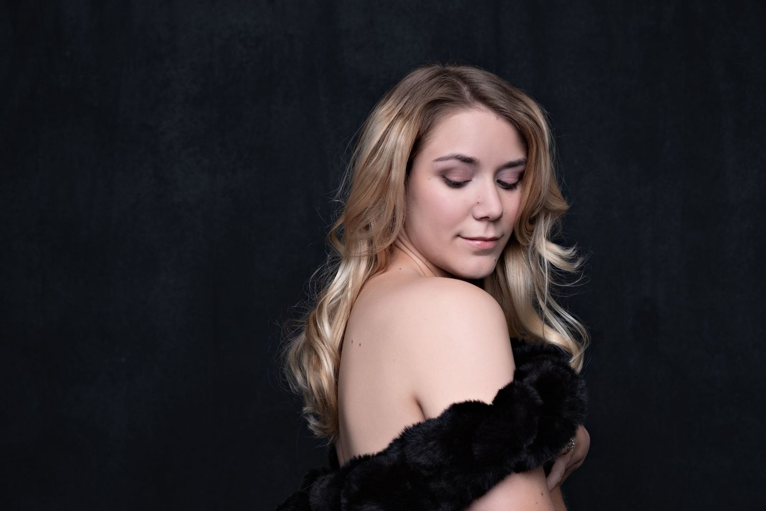 Blonde woman in black floral off-shoulder dress poses elegantly against dark studio background.
