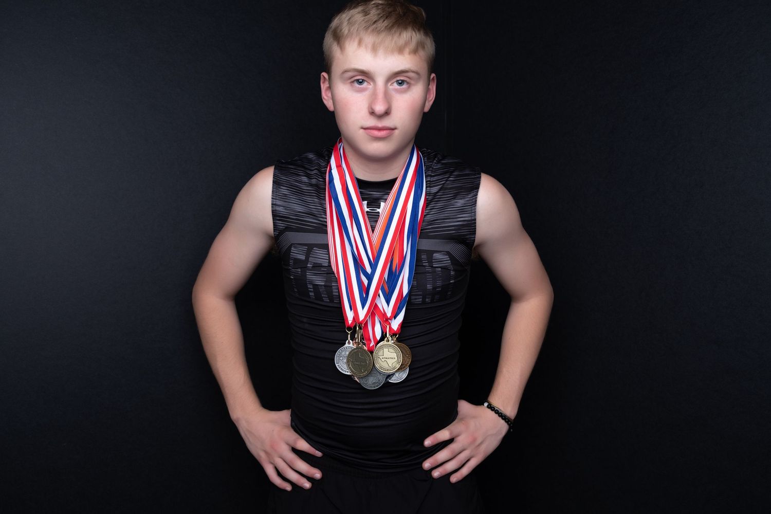 Young male athlete posing with multiple gold and silver medals on red, white, and blue ribbons against dark background.