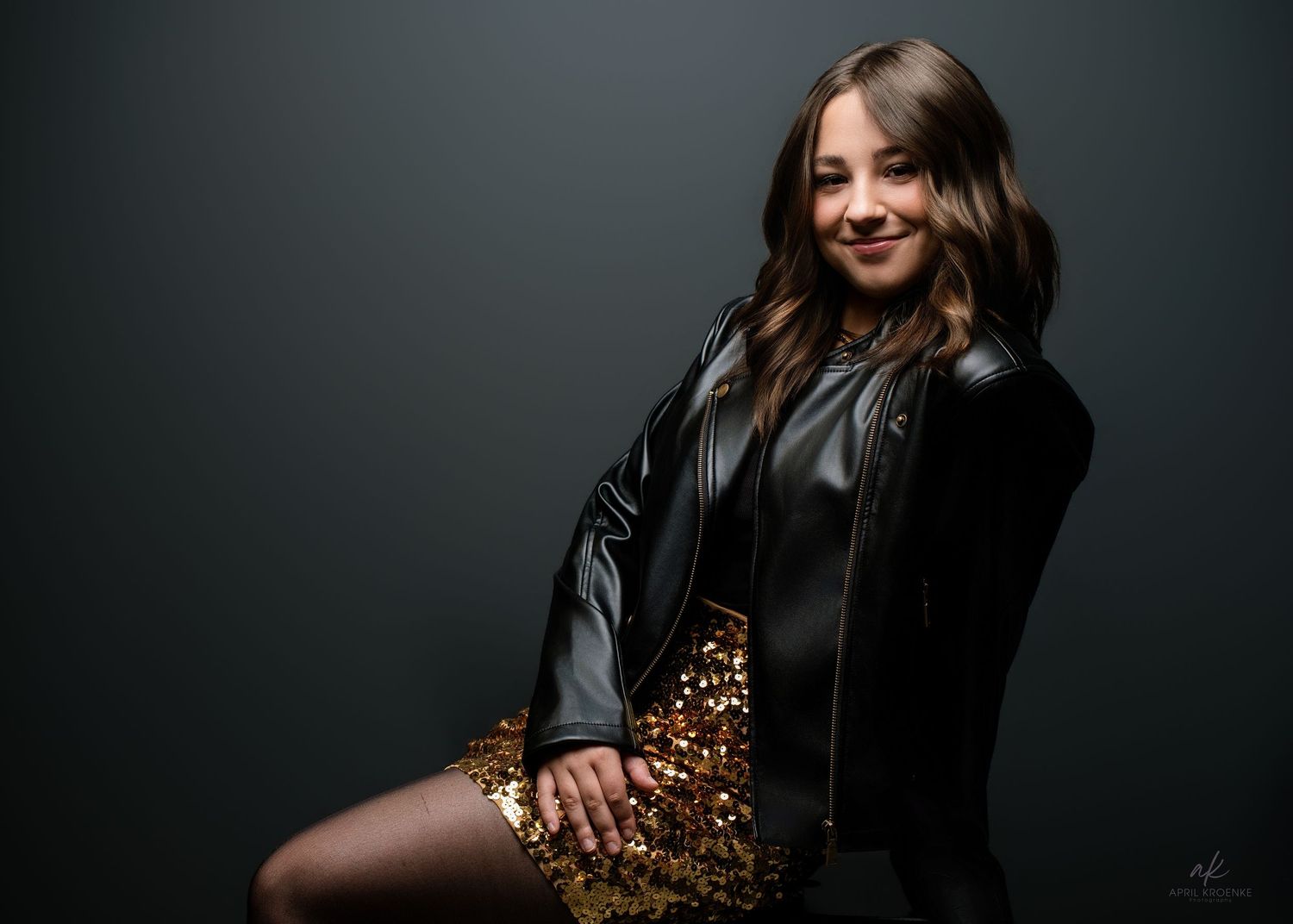 Young woman in sequin skirt and leather jacket poses confidently against dark studio background.