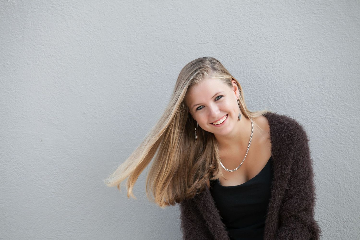 Smiling young blonde woman in a dark cardigan and pearl necklace posing against a light gray wall.