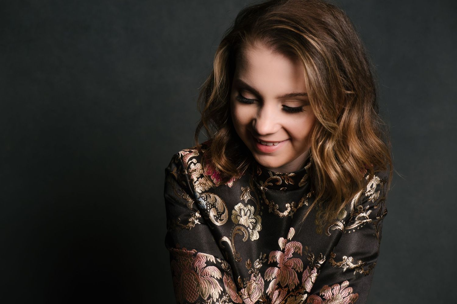 Young woman with wavy brown hair smiles downward, wearing a floral embroidered dress against a dark studio background.