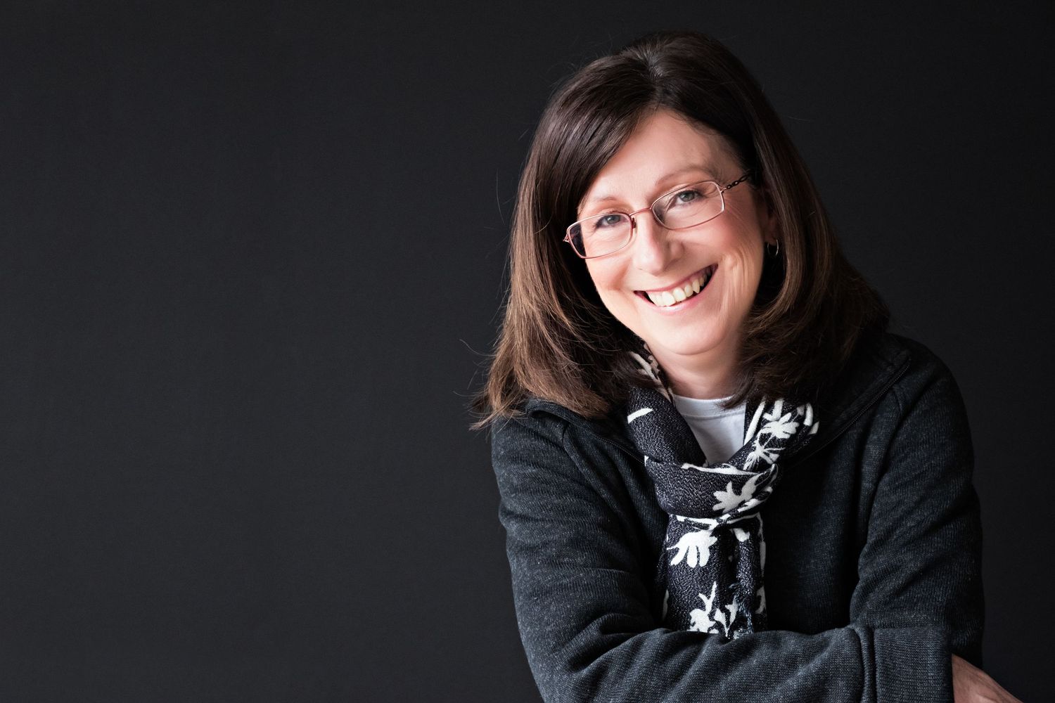 Smiling woman with glasses and brown hair, arms crossed, wearing a dark jacket, posed against a dark background.