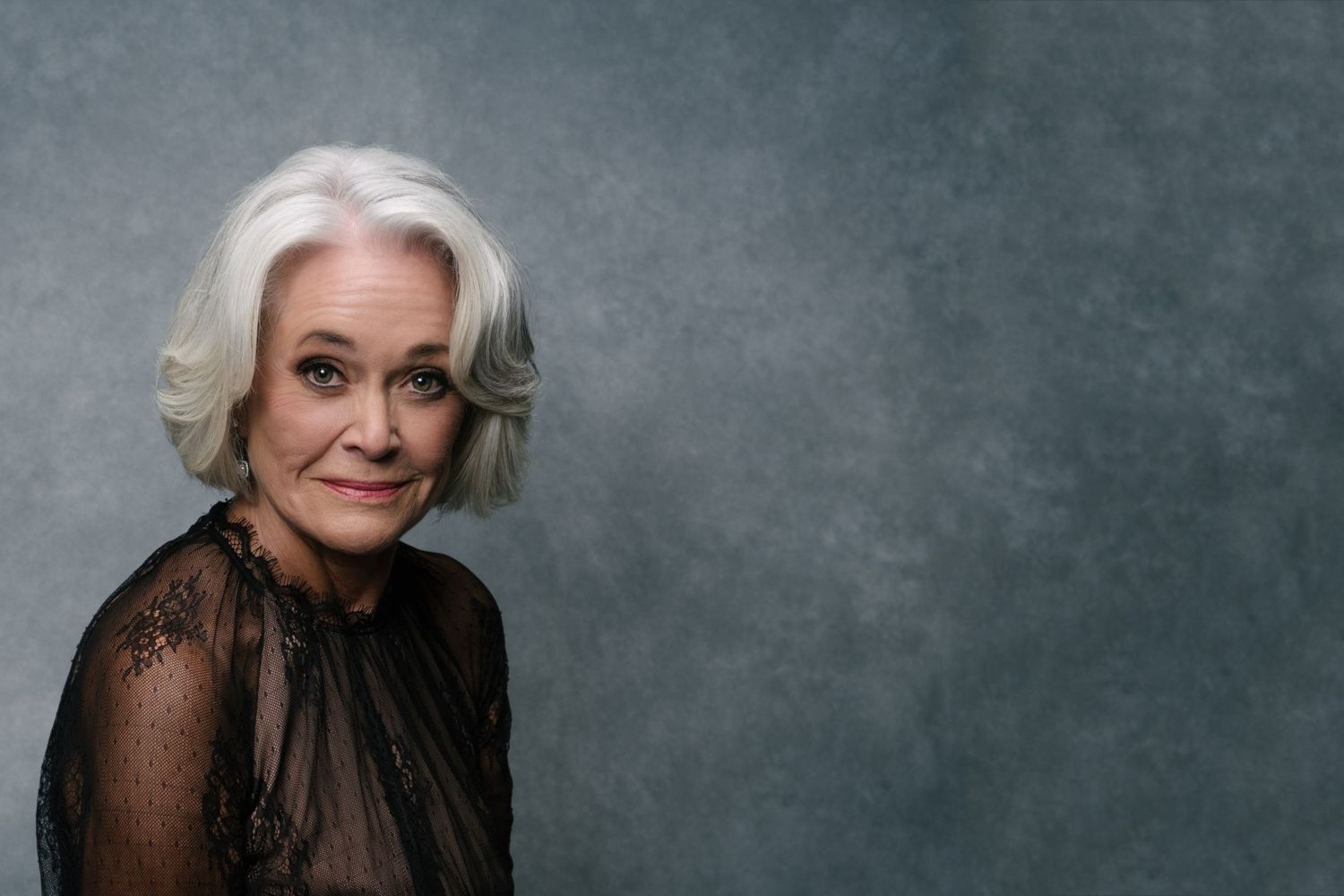 Elegant older woman with silver hair wearing a black lace top, posing against a gray studio background.