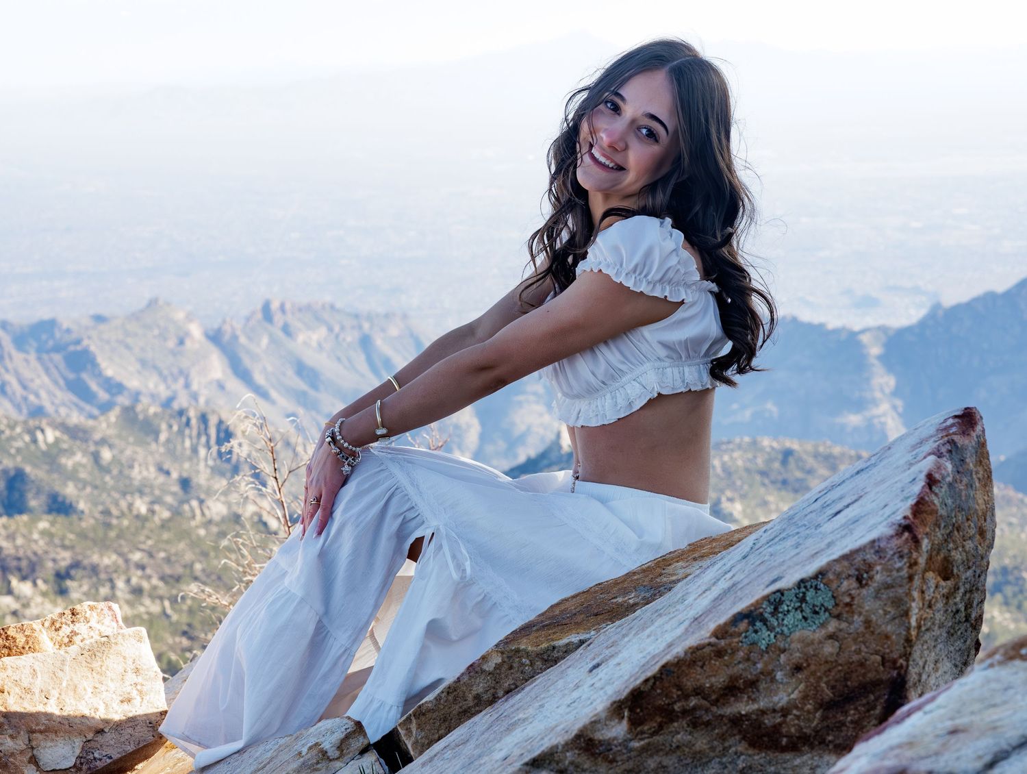 Woman in white crop top and flowy pants sits on rocky mountain summit with scenic mountain range vista in background.