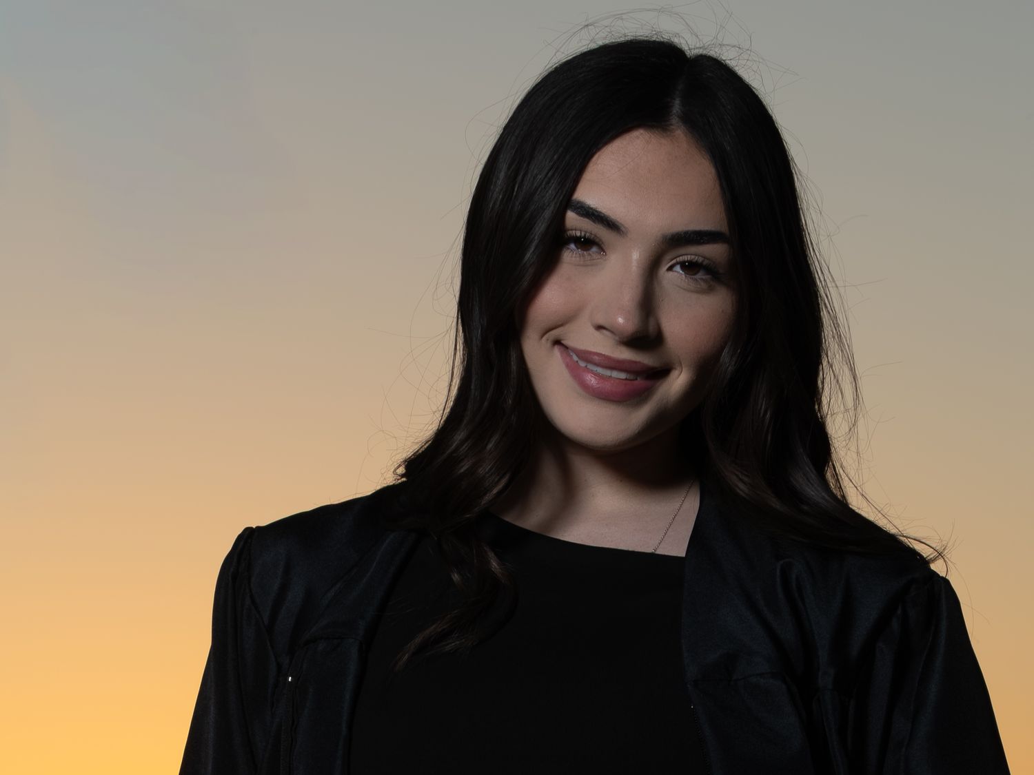 Portrait of a person with long dark hair wearing a black blazer against a sunset sky background.