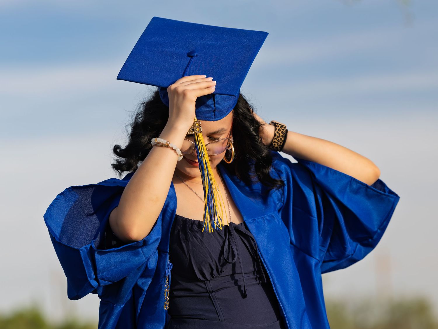 Graduate in blue cap and gown adjusts tassel during outdoor graduation ceremony celebration.
