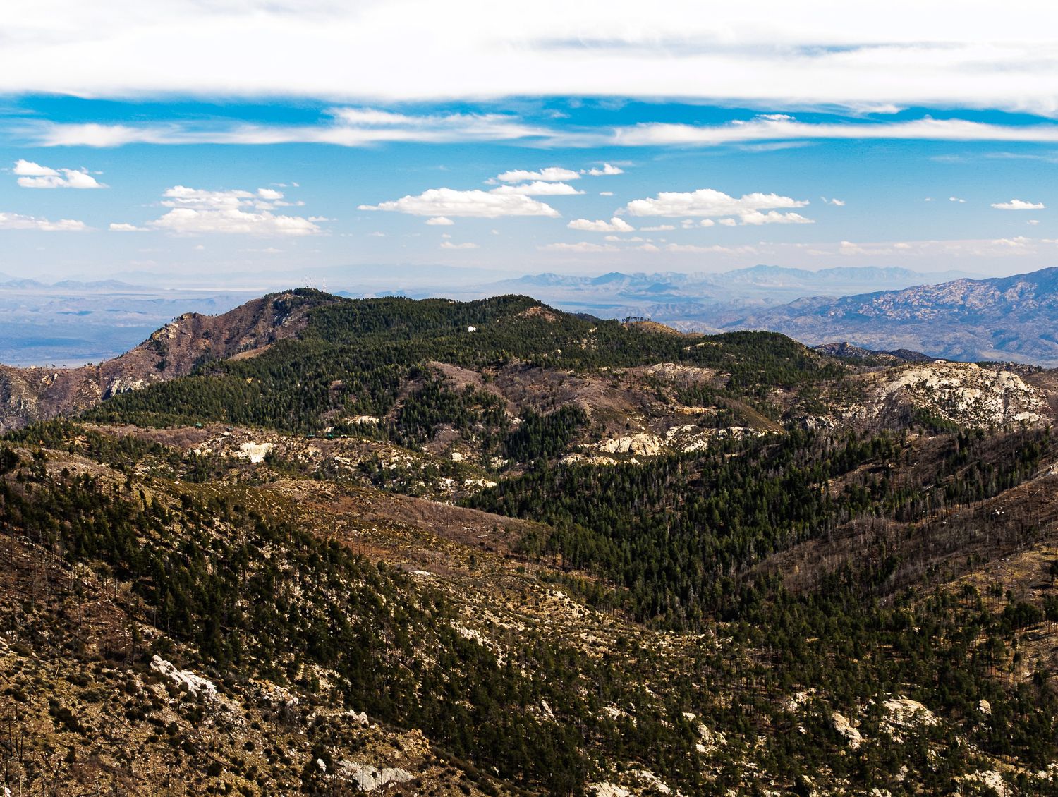 Panoramic mountain landscape with rugged peaks and valleys stretches across the horizon under a bright blue sky.